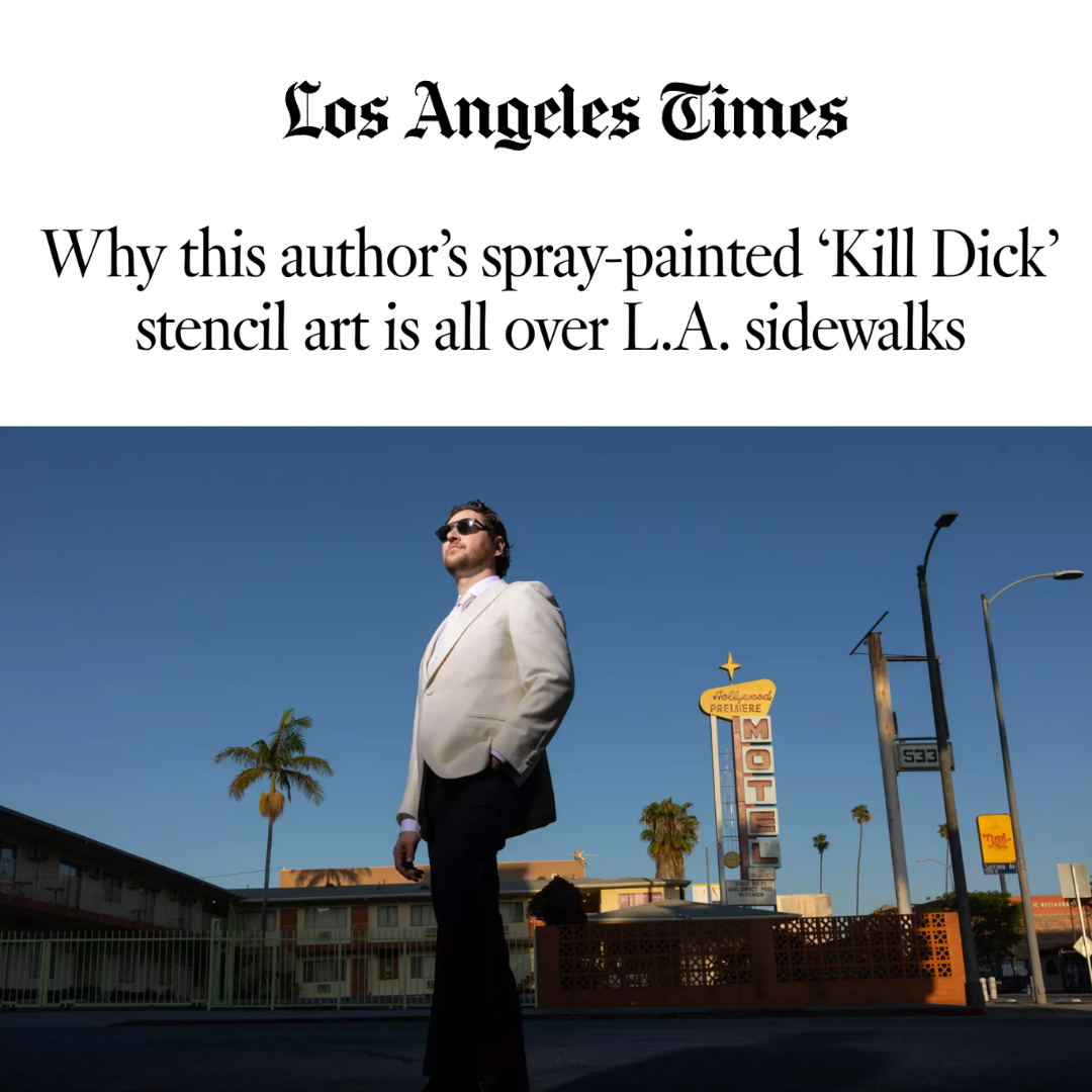 A man in a white suit jacket and sunglasses standing on an LA sidewalk with palm trees, motel, and street signs in the background during the daytime.