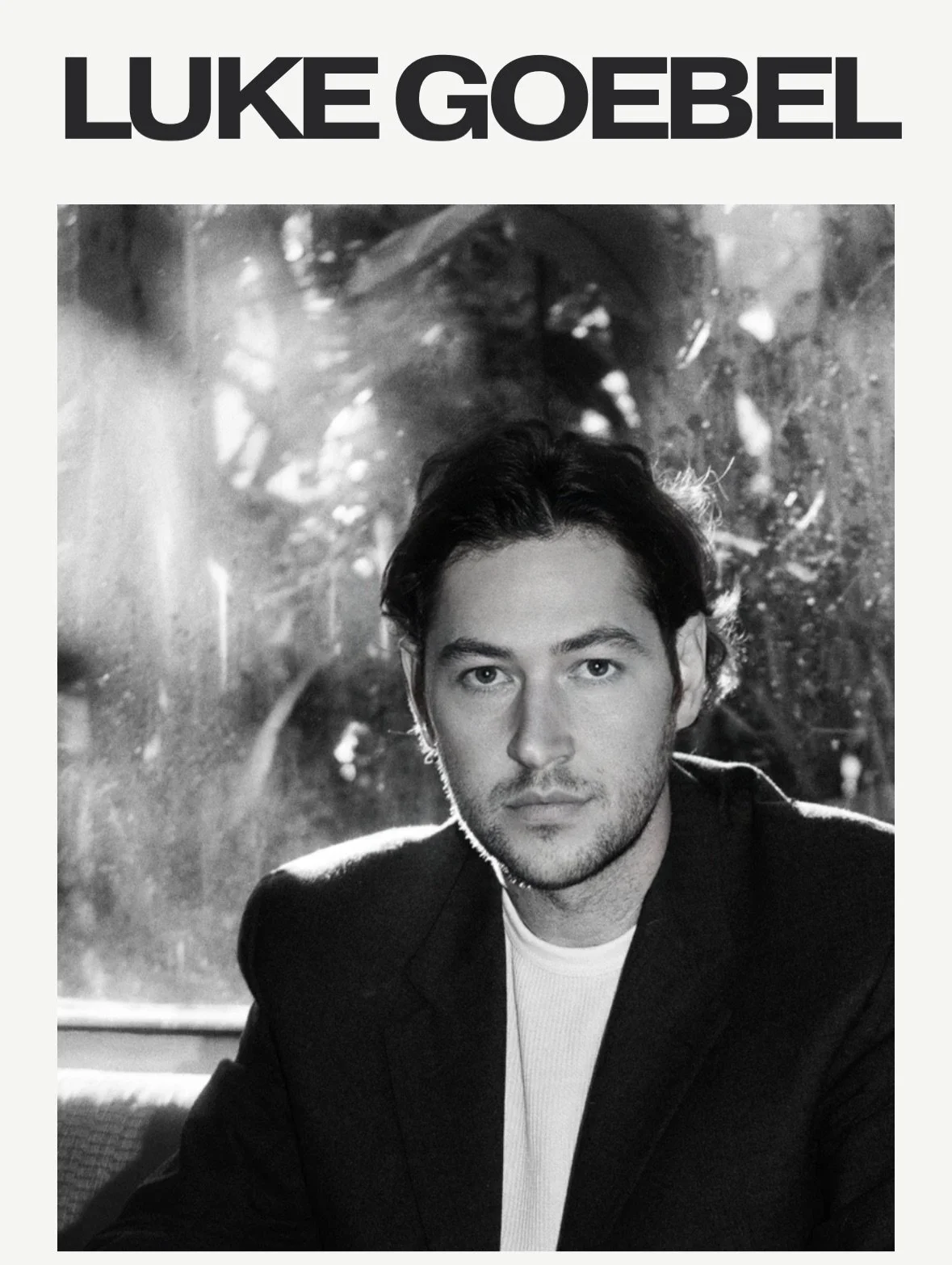 Black and white promotional image of Luke Goebel with his name written in large black letters at the top, featuring a portrait of a young man with dark hair, seated indoors, with a window and trees outside visible in the background.