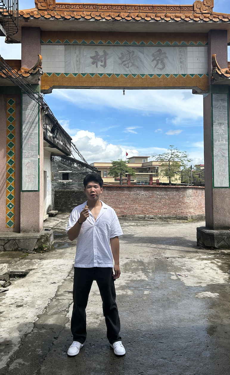 A young man in a white shirt and black pants standing outdoors under a traditional Asian-style archway with Chinese characters, holding a small object in his right hand, with a brick wall and buildings in the background and a partly cloudy sky.