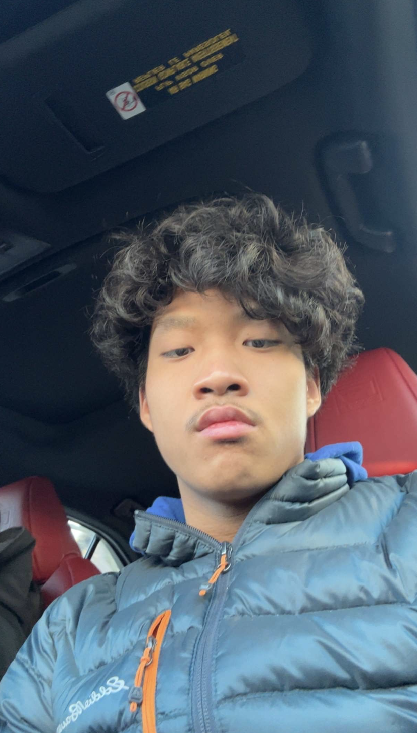 A young man with curly hair taking a selfie inside a car with red seats, wearing a blue puffer jacket.