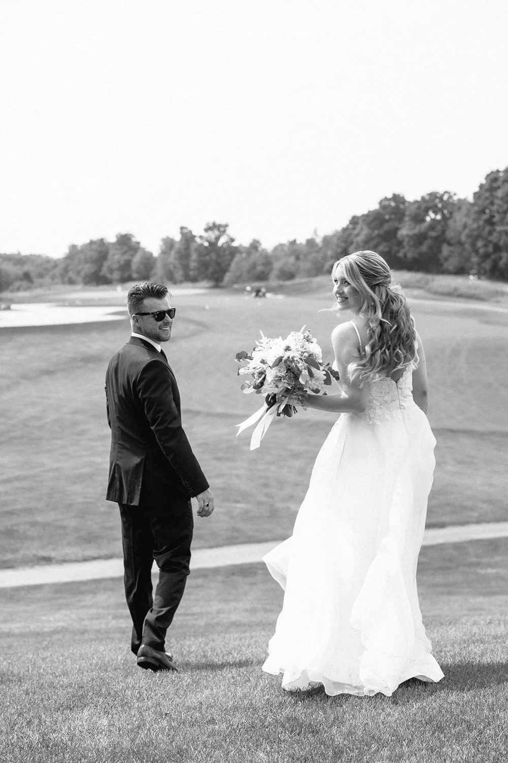 A bride in a wedding dress holding a bouquet standing on golf course smiling at a groom 