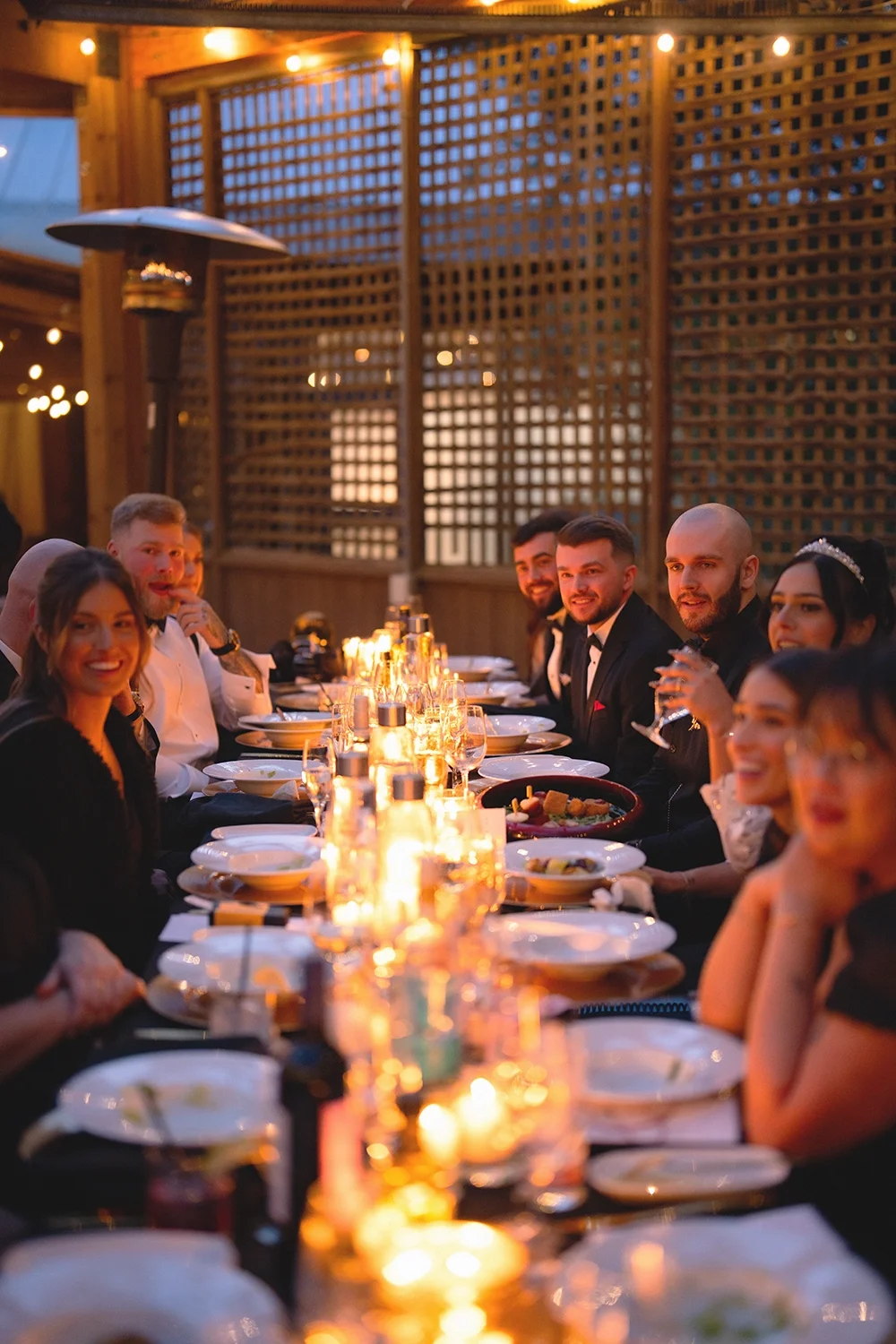 People dressed in formal attire sitting at a long dinner table with candles, plates, and glasses, enjoying an outdoor evening wedding