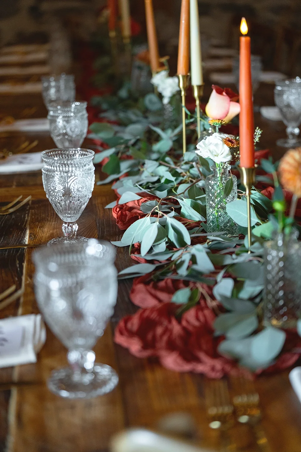 Elegant table setting with glass goblets, a floral centerpiece with white and pink flowers, green foliage, tall orange candles, and gold accents.
