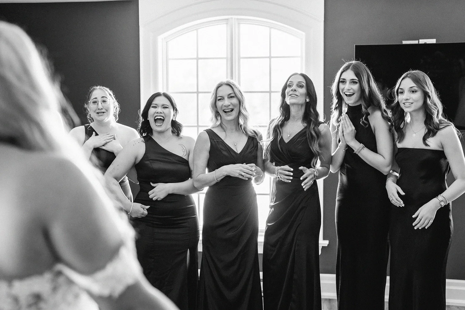 Six bridesmaids in black dresses laughing and smiling at the bride reveal 