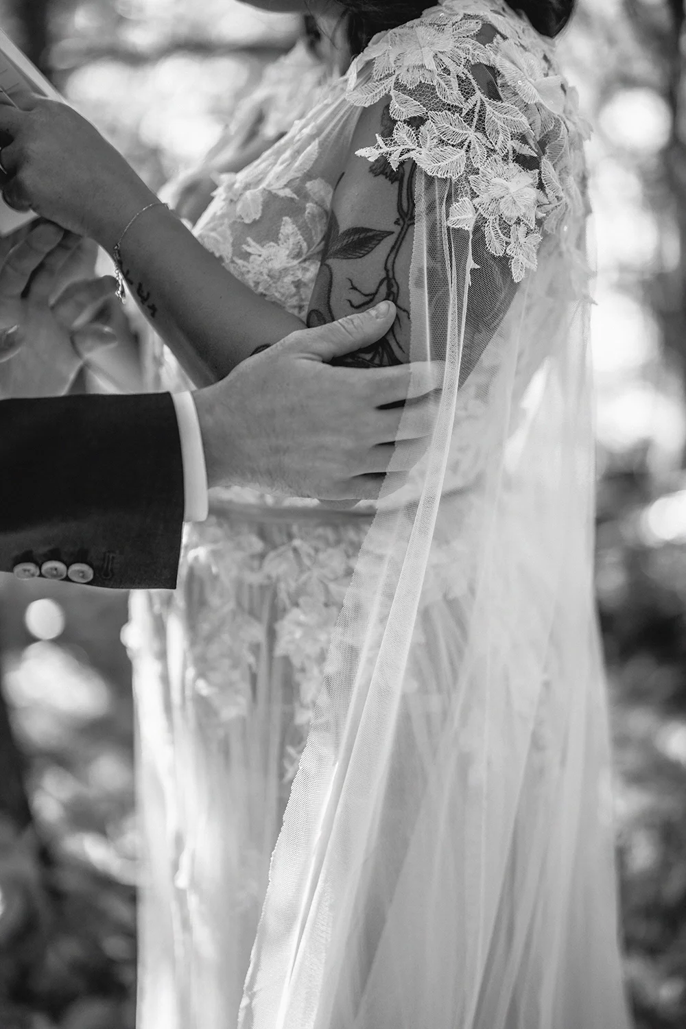 A bride and groom hold hands during their wedding ceremony, with the groom placing his hand on the bride's arm. The bride is wearing a lace wedding dress with floral embroidery, and the groom is in a suit. The photo is in black and white.
