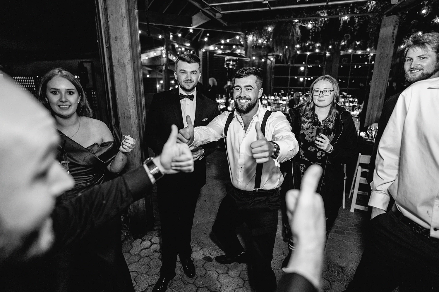 Group of people at a wedding reception, some giving thumbs up, in a decorated venue at night.