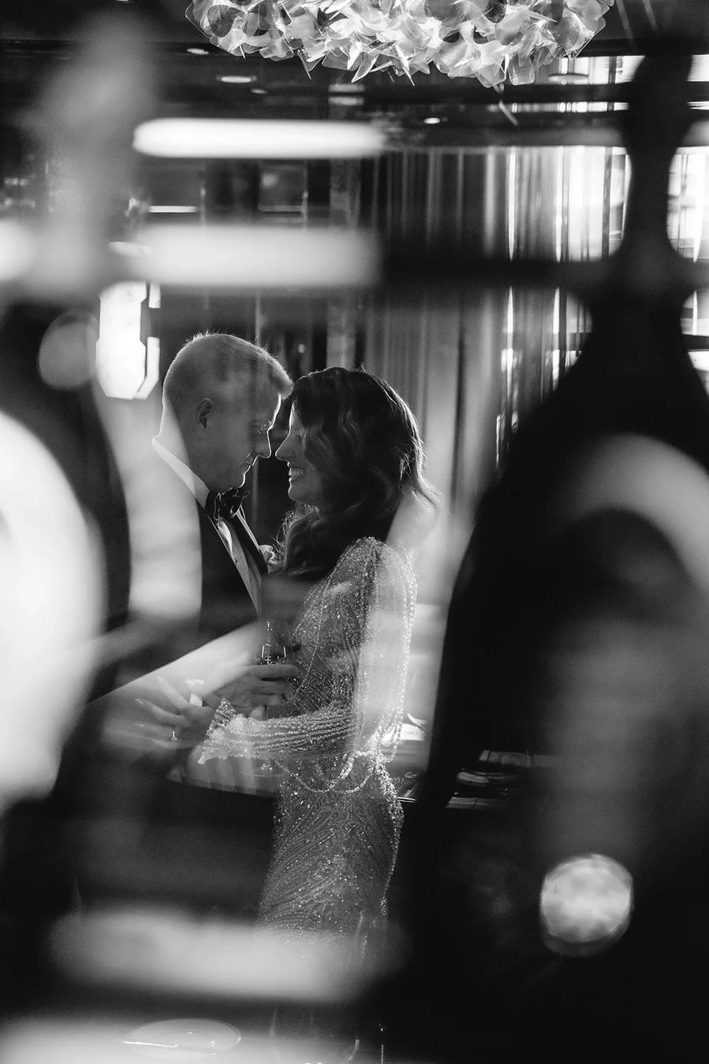 Black and white photo of a woman and man in formal attire sharing a close moment, holding glasses, seen through a decorative framework.