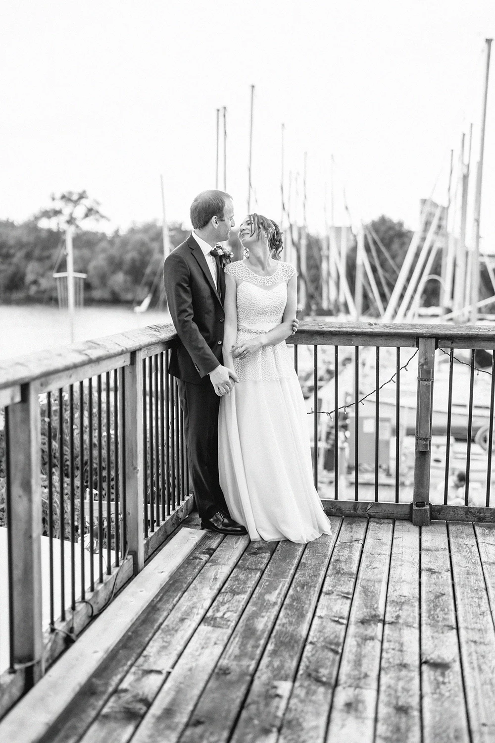 A black and white photo of a bride and groom standing on a wooden deck near sailboats, looking at each other lovingly.