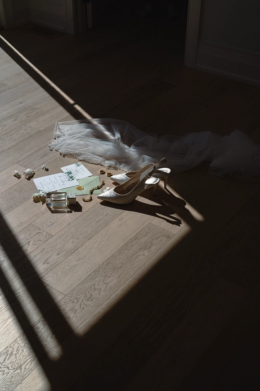 A collection of wedding items on a wooden floor illuminated by sunlight, including a white wedding dress, a pair of white high-heeled shoes, wedding invitations, a small glass bottle, a green envelope, a necklace, and some small decorative items.