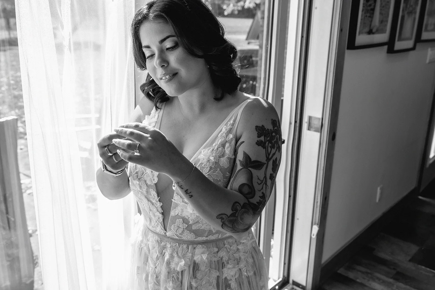 A woman in a lace wedding dress standing indoors near a window, holding her hands together, with tattoos on her arm, smiling softly.