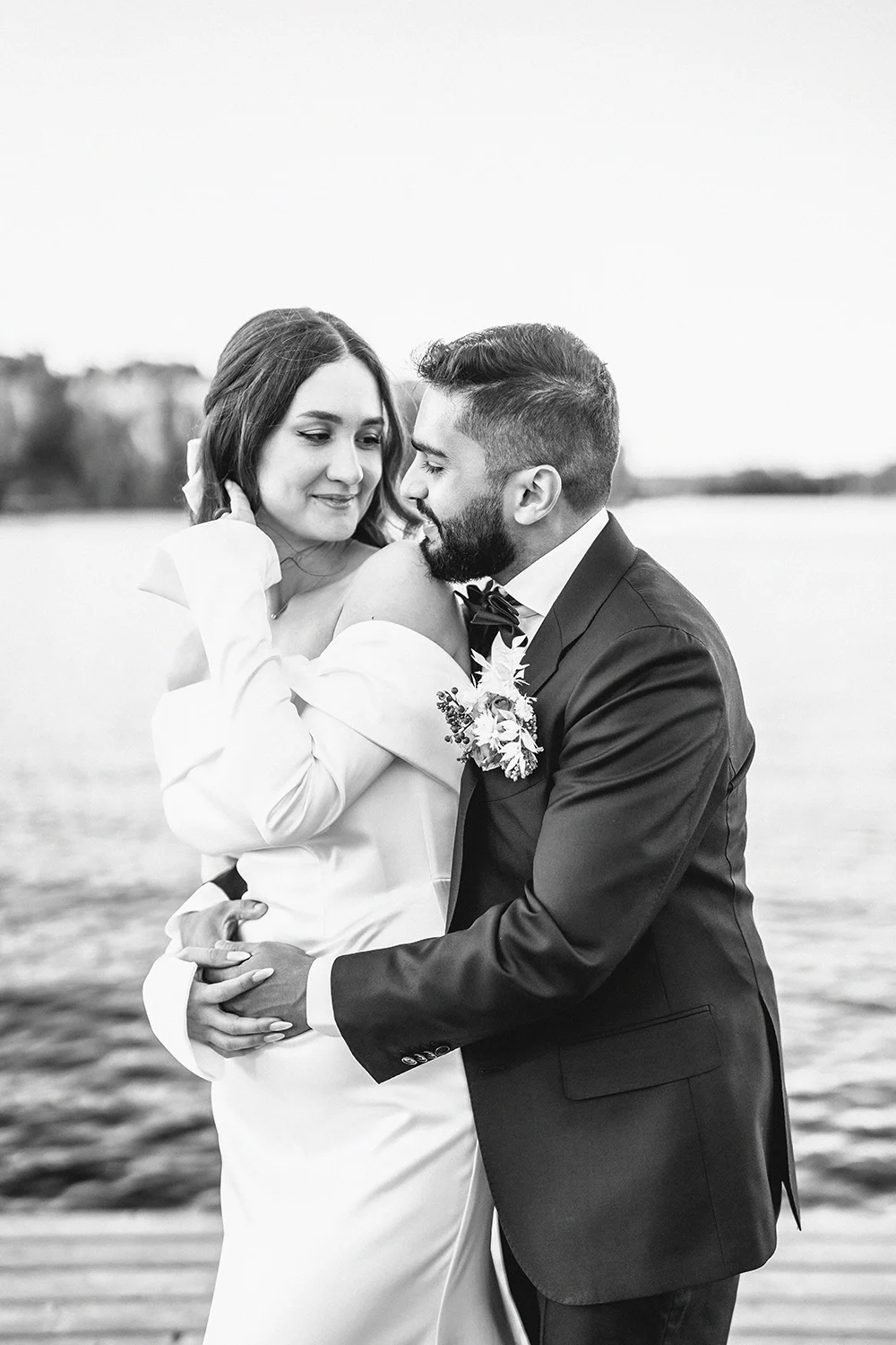 Black and white photo of a bride and groom embracing by a body of water, with the bride wearing an off-the-shoulder wedding dress and the groom in a suit with a bow tie.