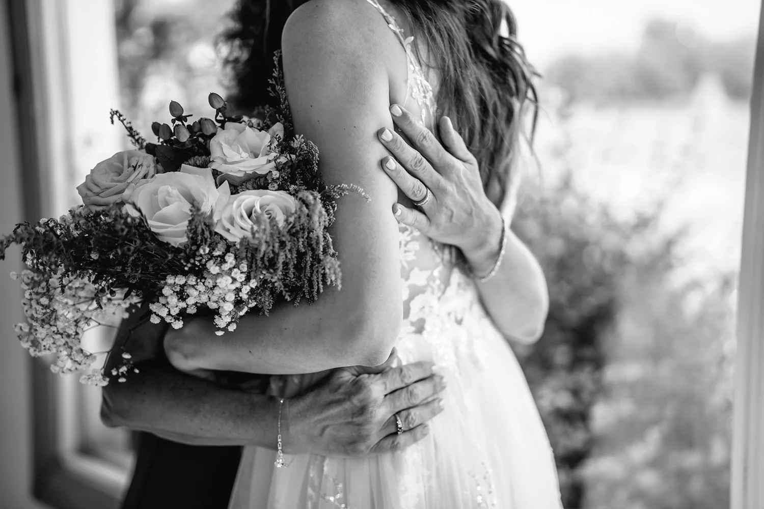 A woman in a wedding dress holding a bouquet of roses and flowers, with her hands on her chest, sharing a close embrace with another person, in front of a window.