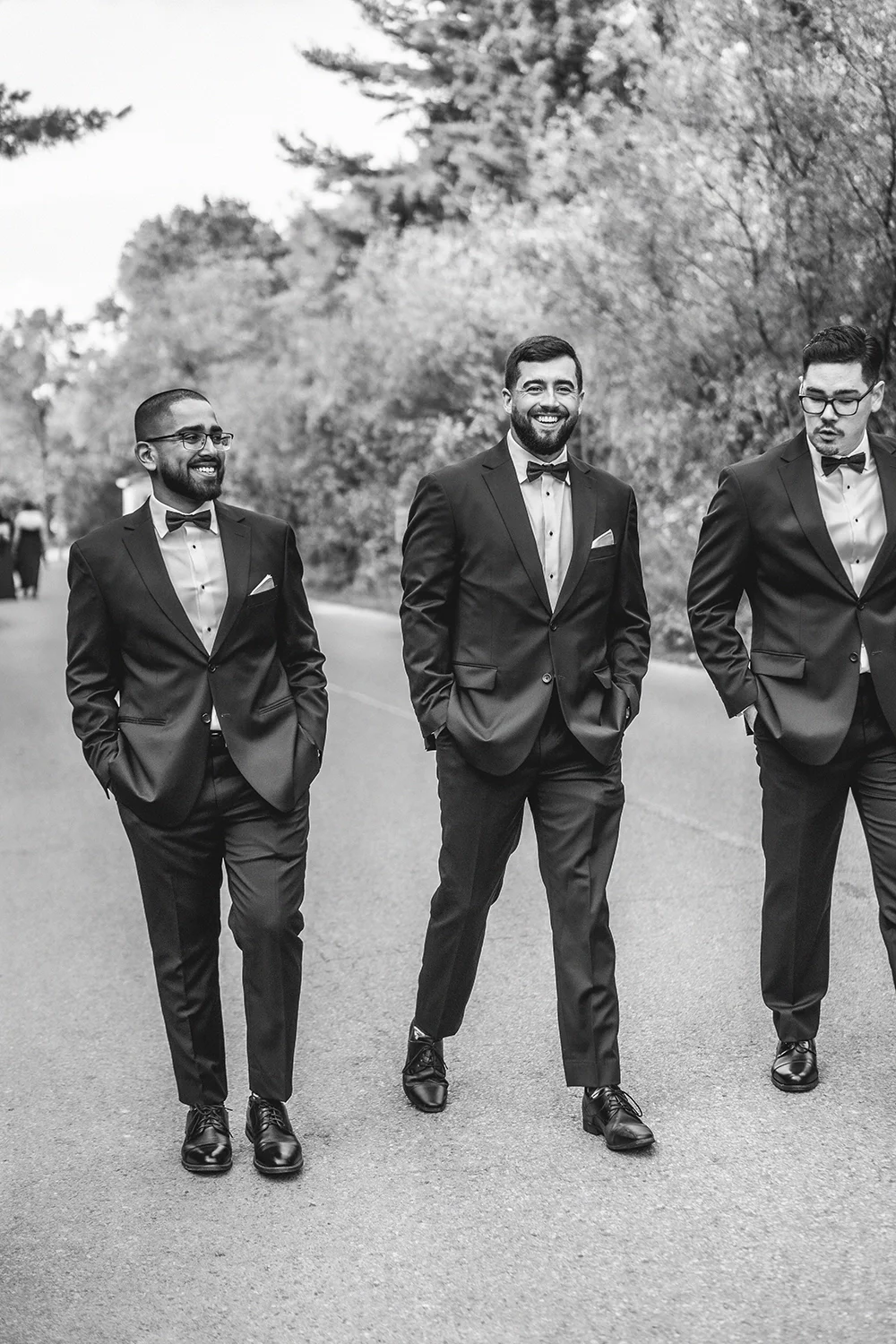 Three men in tuxedos walking on a road outdoors, smiling, with trees in the background.