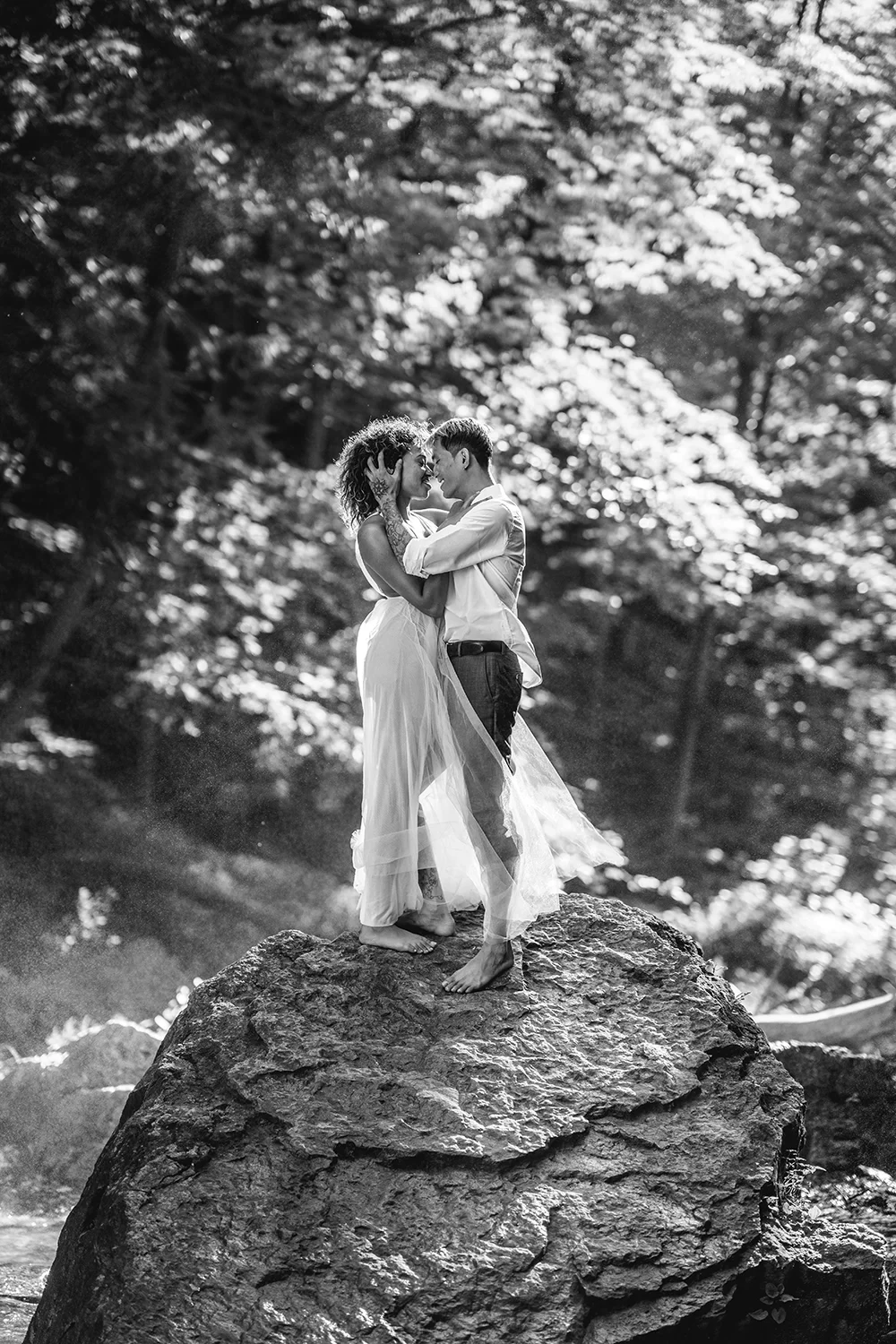A couple standing on a large rock in a forest, embracing and touching foreheads, with sunlight filtering through trees in the background.