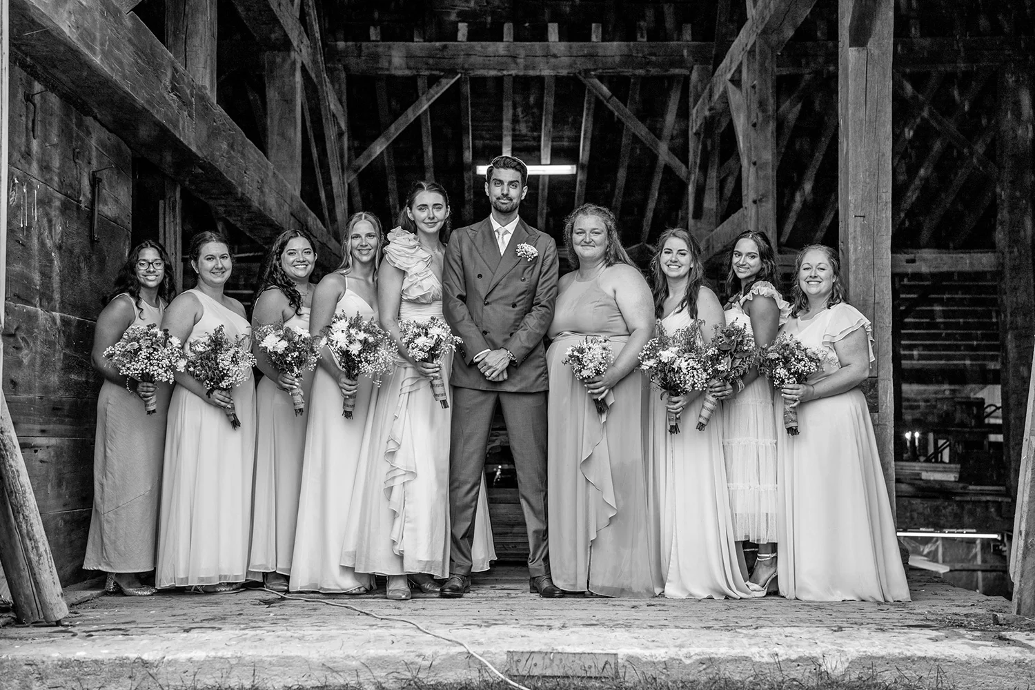 Black and white photo of a wedding party standing in a rustic barn. The group includes a groom in a double-breasted suit and nine bridesmaids in long dresses, all holding bouquets of flowers.