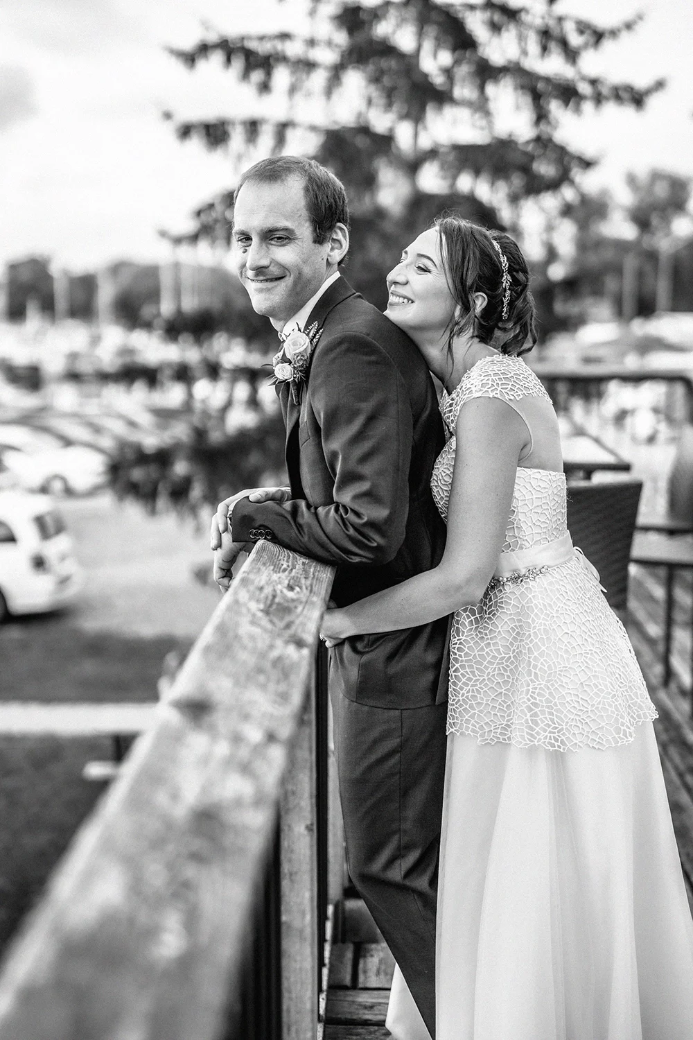 Black and white photo of a bride and groom leaning on a wooden railing, smiling and enjoying a moment together outdoors during their wedding.