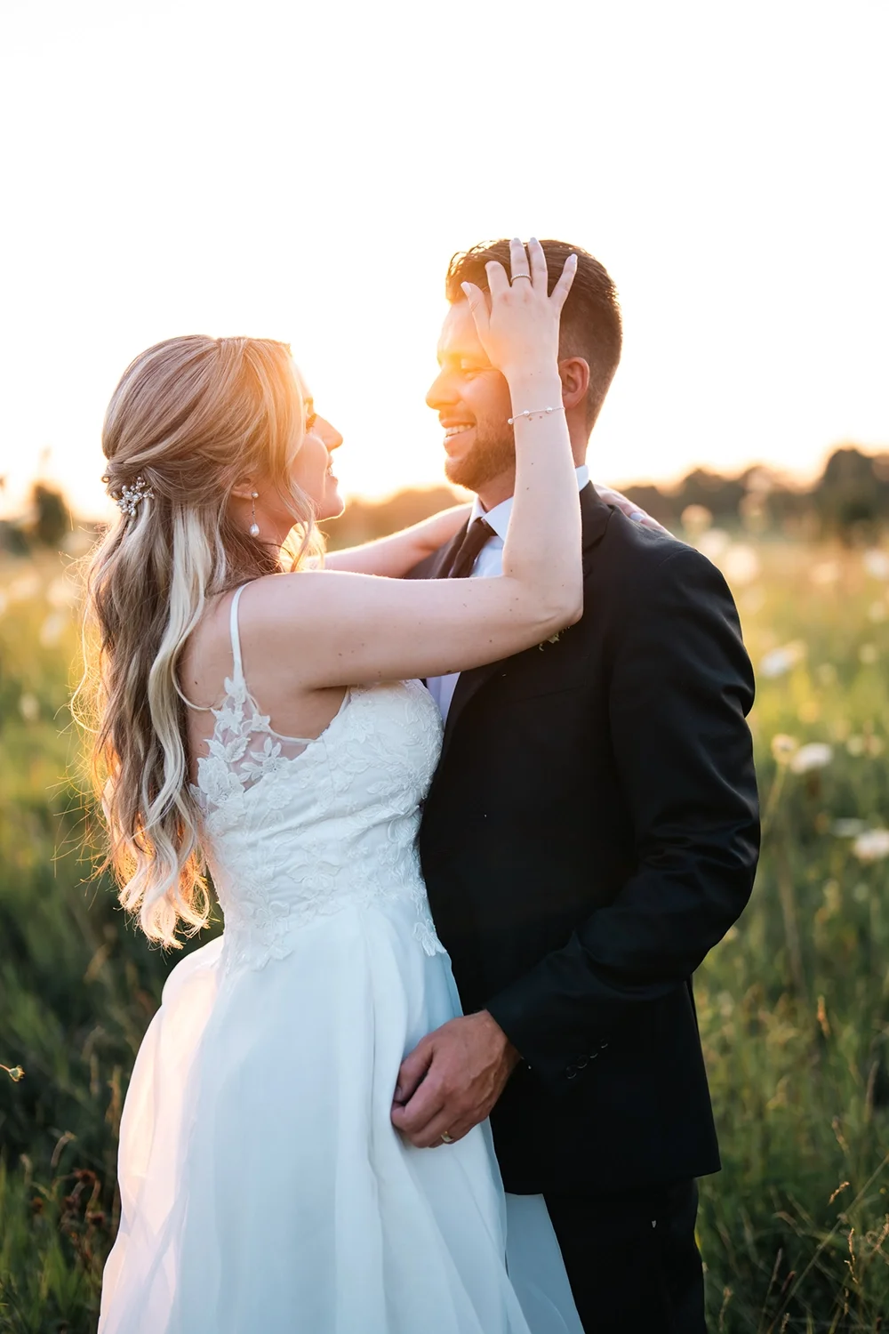 A bride and groom sharing a moment in a field at sunset, smiling and embracing each other.