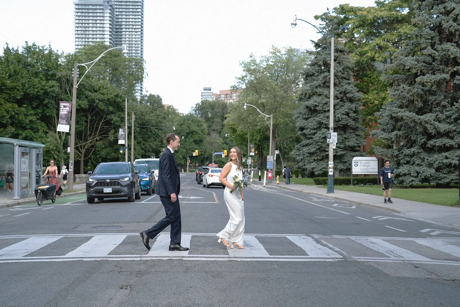 A bride and groom crossing a city street on a crosswalk, with the bride holding a bouquet of flowers, in an urban park area with trees, cars in the background, and a few pedestrians. Toronto university 
