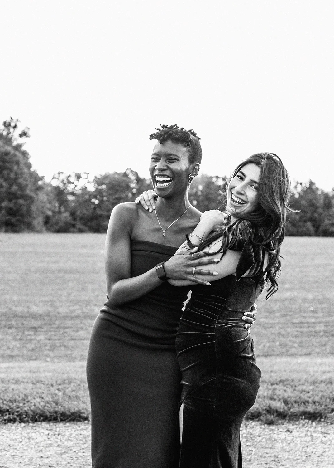 Two women wedding guests laughing and hugging outdoors in a grassy field with trees in the background, wearing elegant dresses.
