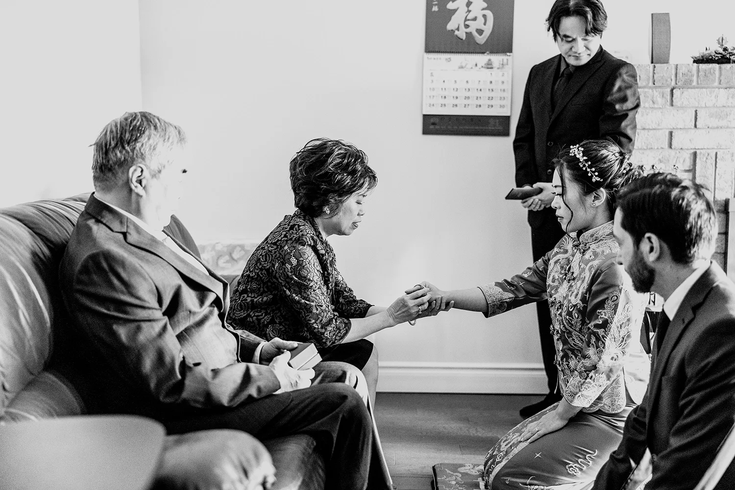A woman in traditional Asian attire holding hands with an older woman, with three other people observing in a living room, during a formal or cultural ceremony.