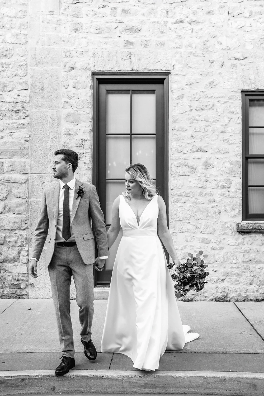A black-and-white photo of a bride and groom holding hands, walking outside a stone building with large windows.