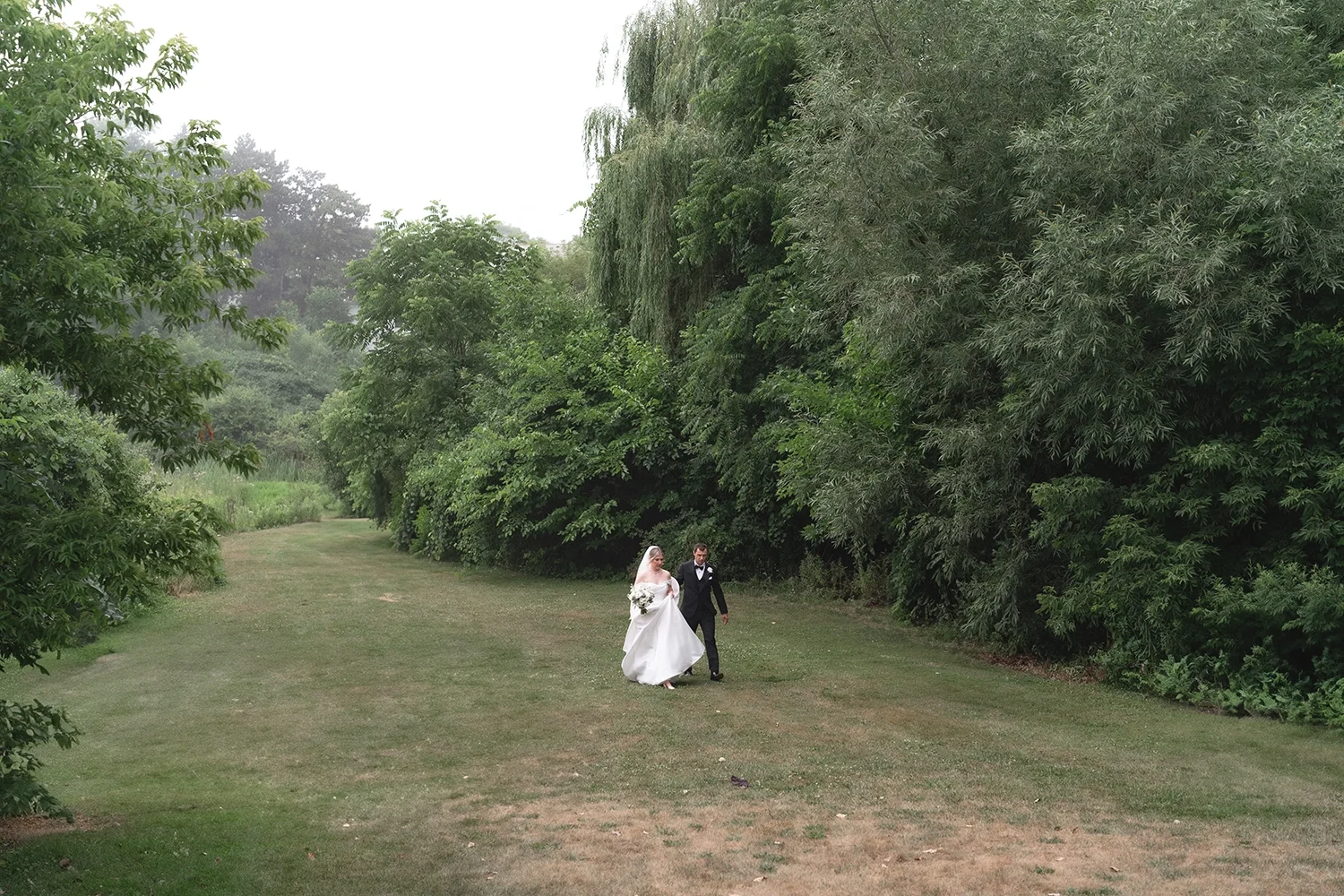 A bride in a white wedding gown and veil holding a bouquet, walking arm-in-arm with a groom in a black tuxedo on a grassy path surrounded by lush green trees.