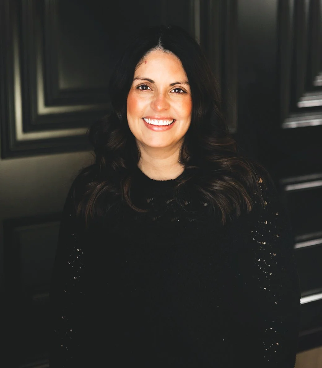 A woman with long dark hair, smiling and wearing a black sweater, sitting in front of a dark wood-paneled wall.