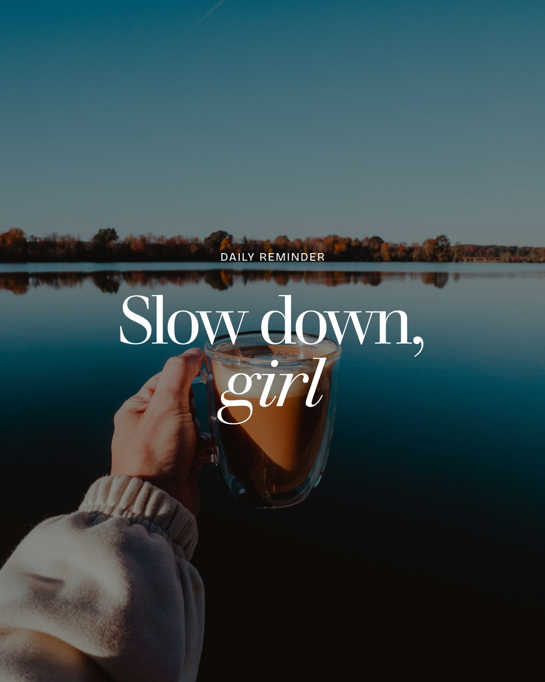 A person holds a glass mug filled with coffee or tea, with a scenic lake and trees in the background, displaying a motivational message to slow down, girl.