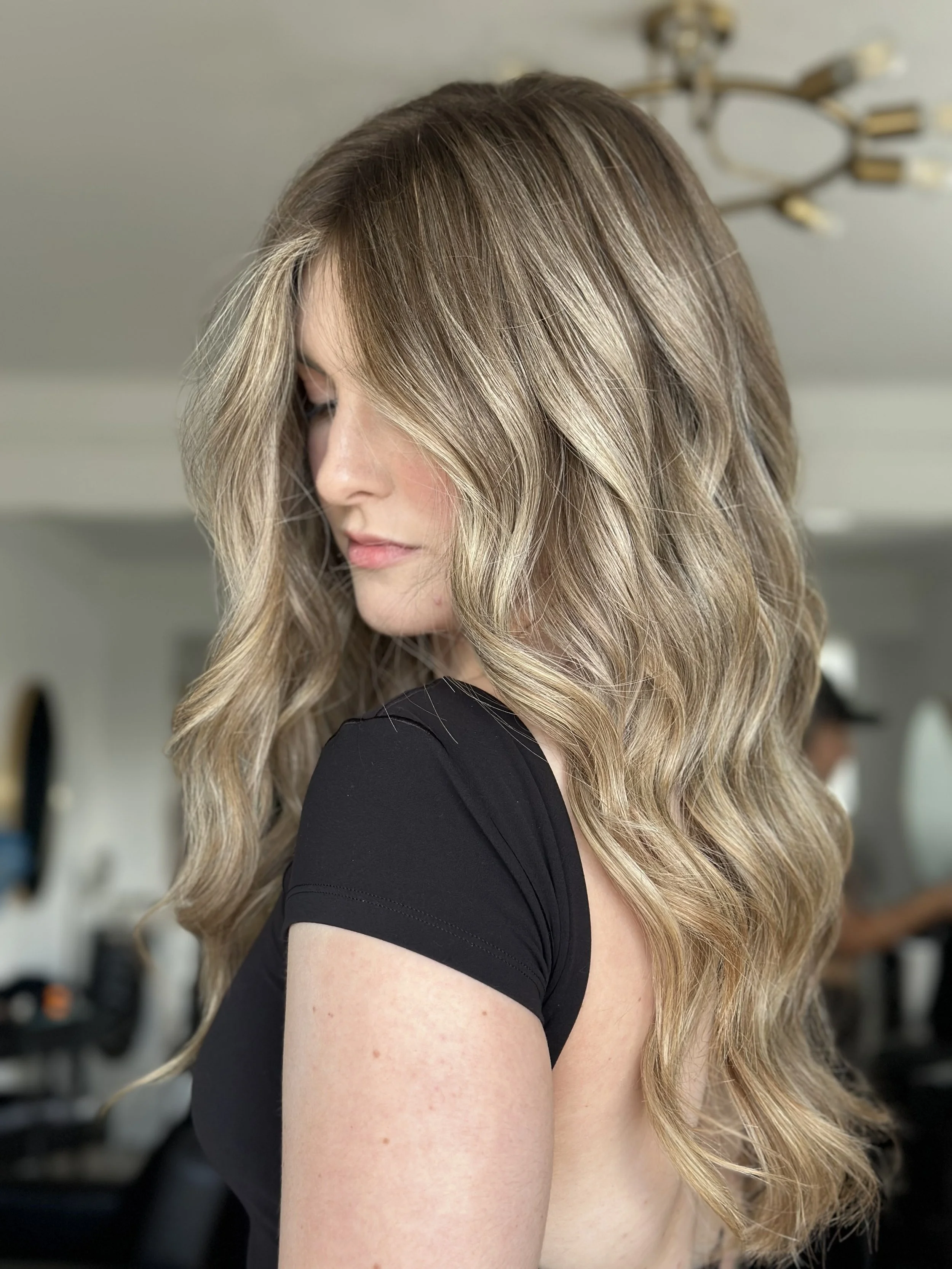 A woman with long wavy blonde hair and light skin, looking down, wearing a black top, in a room with modern decor.
