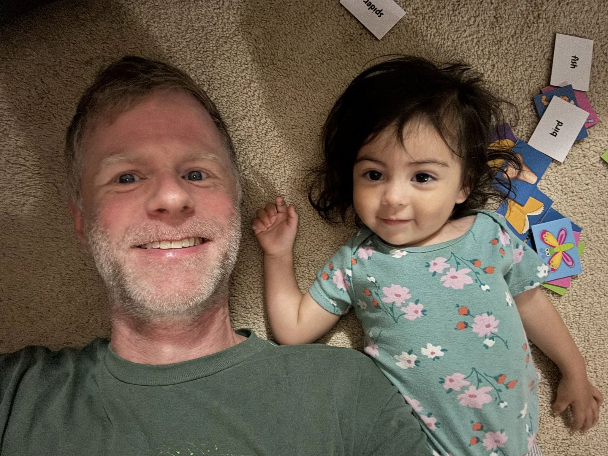 A smiling man with gray hair and a beard lying on a beige carpet next to his daughter with dark hair wearing a teal floral dress, surrounded by scattered word and picture cards.