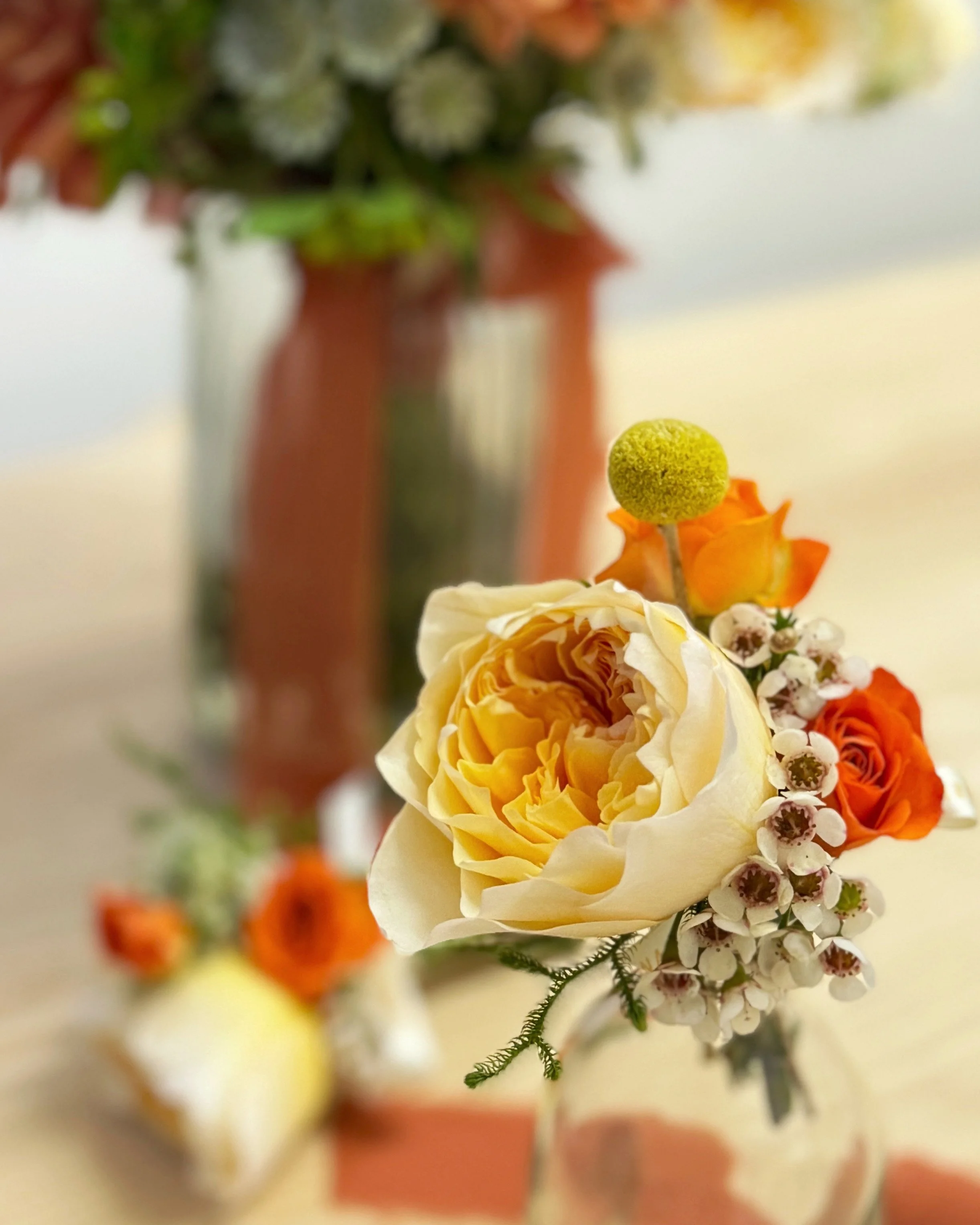 Close-up of a floral arrangement featuring a large, cream-colored rose, orange roses, white small flowers, and a round green berry in a glass vase with a blurred background.