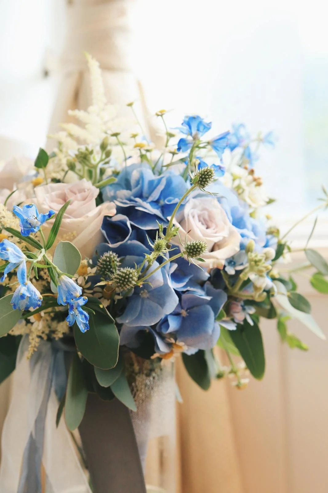 A bouquet of blue, white, and pink flowers with greenery, arranged in a vase, near a window.