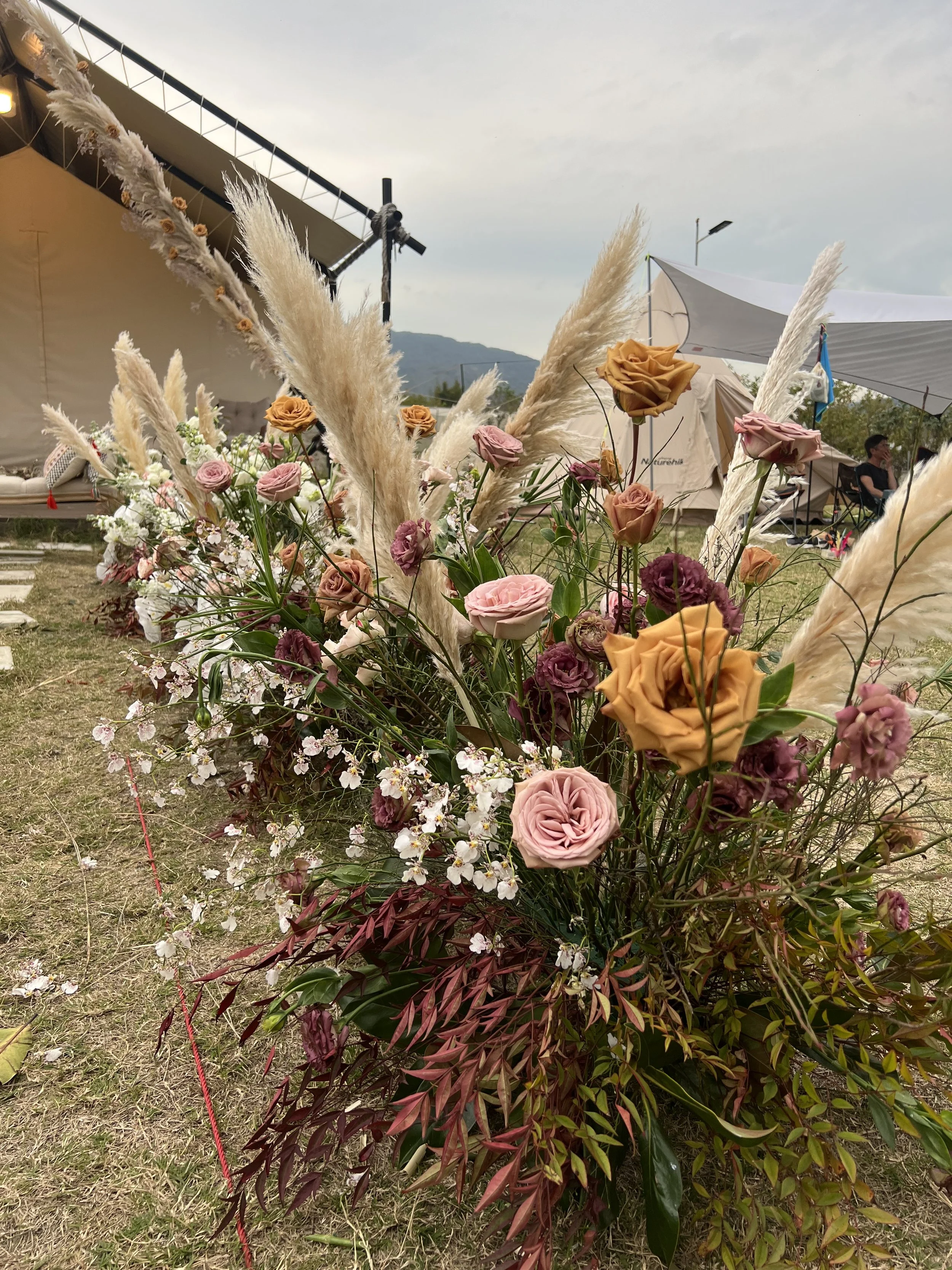 A colorful floral arrangement with blush, peach, and purple roses, white and pink blossoms, and tall beige pampas grass, set outdoors with tents and cloudy sky in the background.