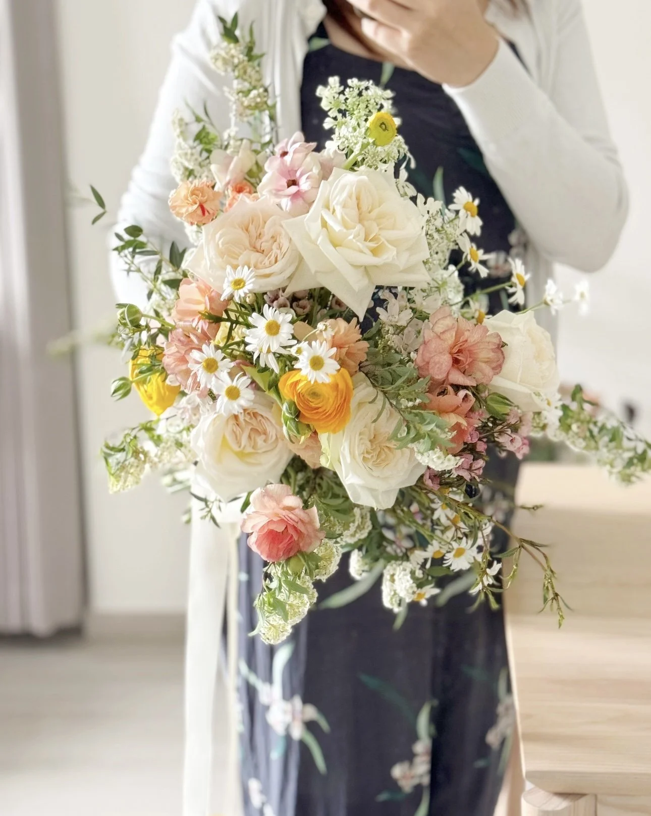 Person holding a large bouquet of assorted flowers including white roses, daisies, pink, yellow, and peach blooms, with a blurred indoor background.