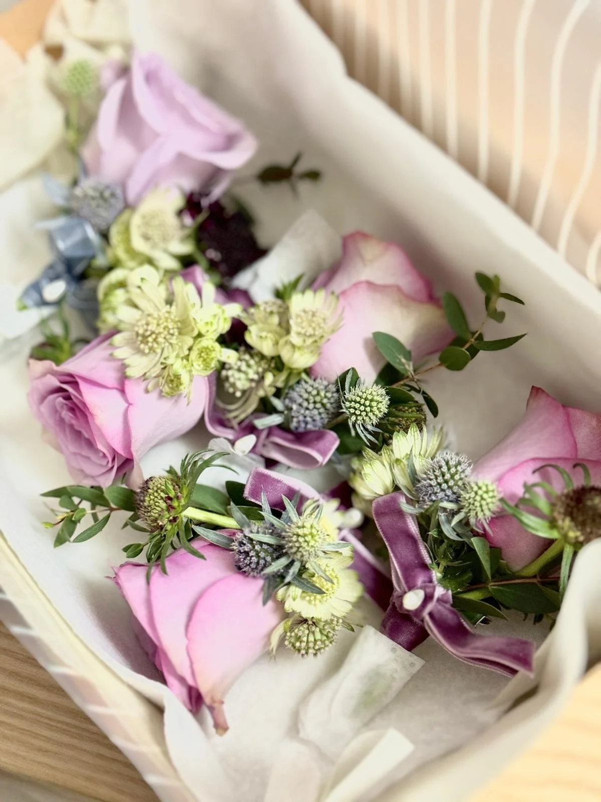 A floral arrangement with pink roses, small white flowers, purple ribbons, and green foliage in a white box.