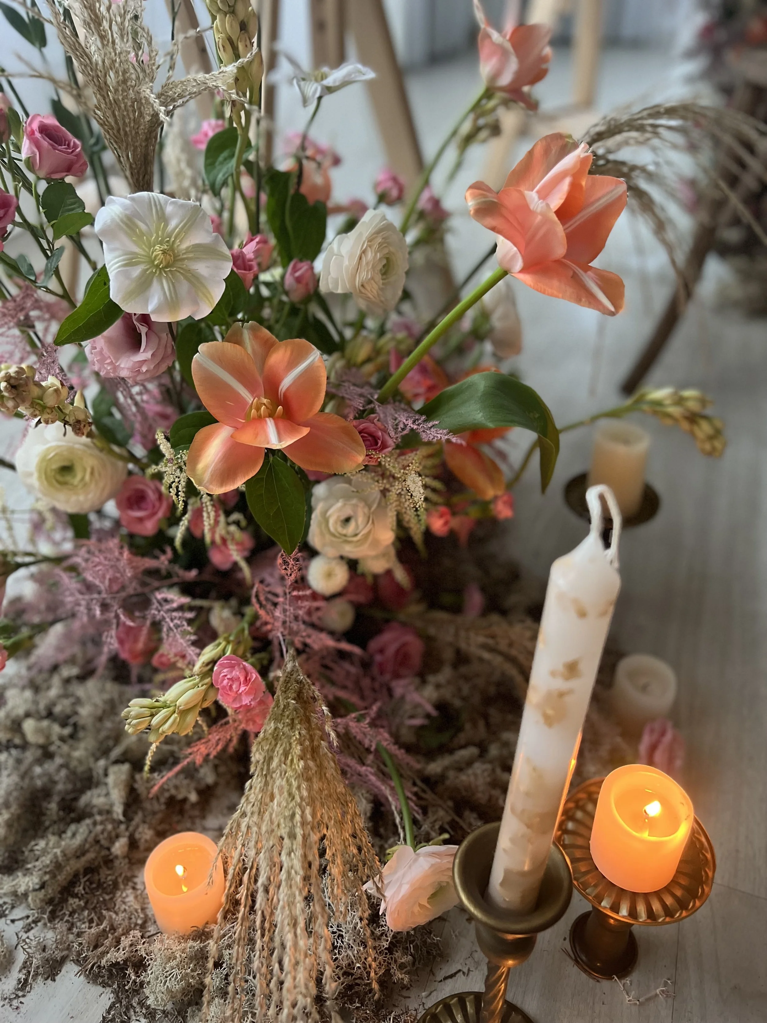 A floral arrangement with pink, white, and orange flowers, surrounded by candles and candles holders, on a table.