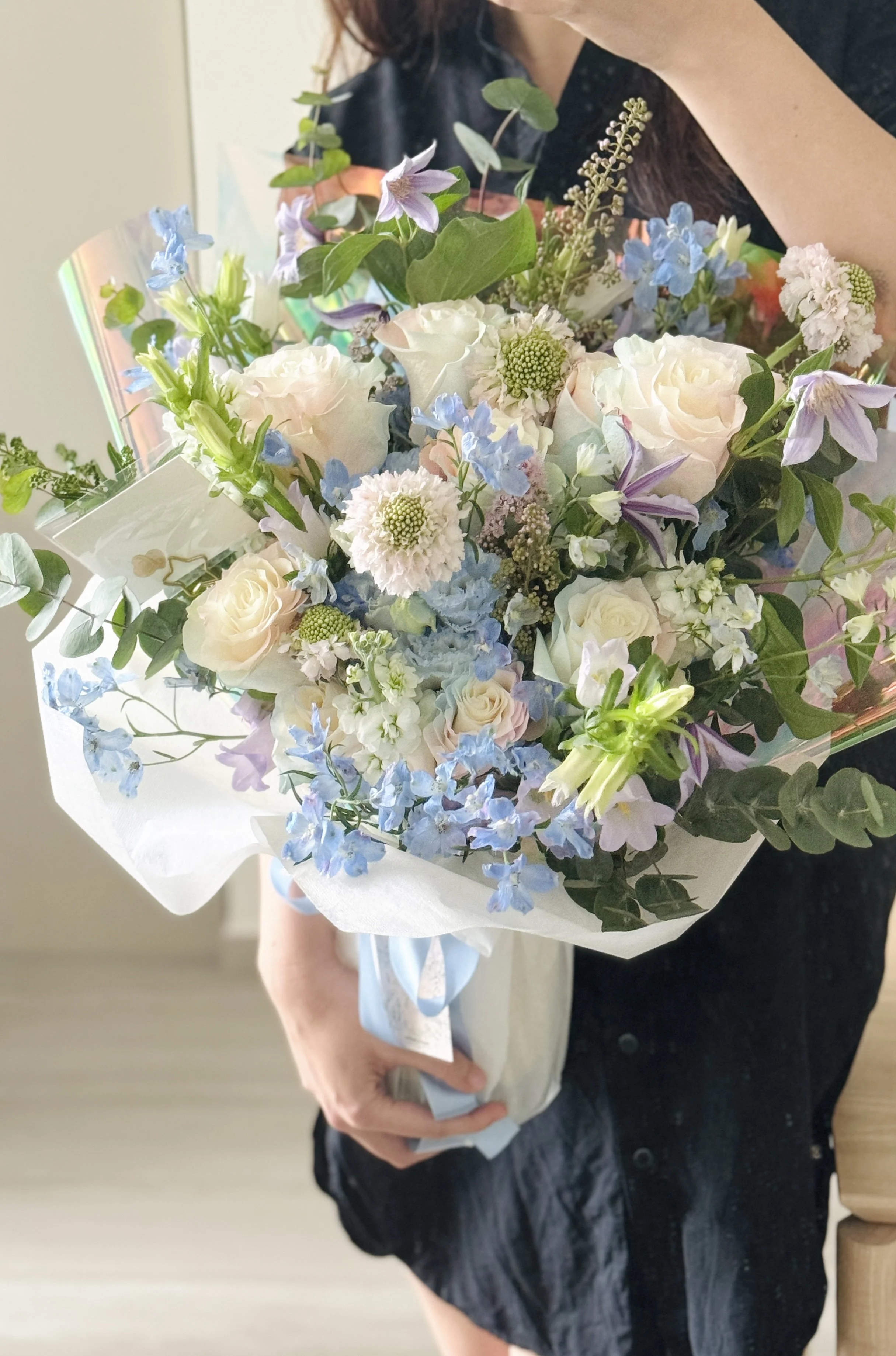 A person holding a bouquet of mixed flowers including white roses, blue delphiniums, and other light-colored blooms wrapped in white paper.