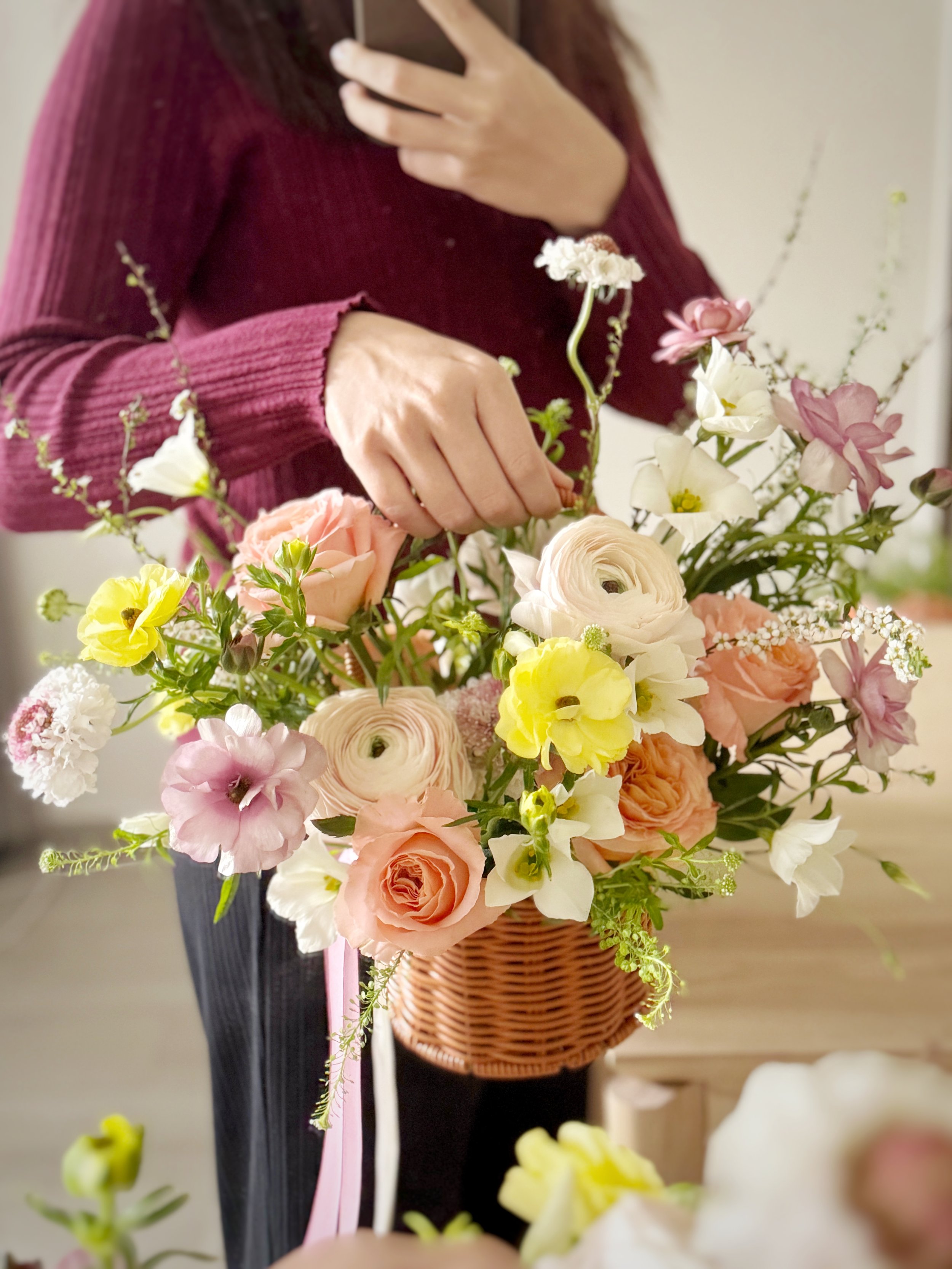 Person in a maroon sweater arranging a colorful flower bouquet in a wicker basket.