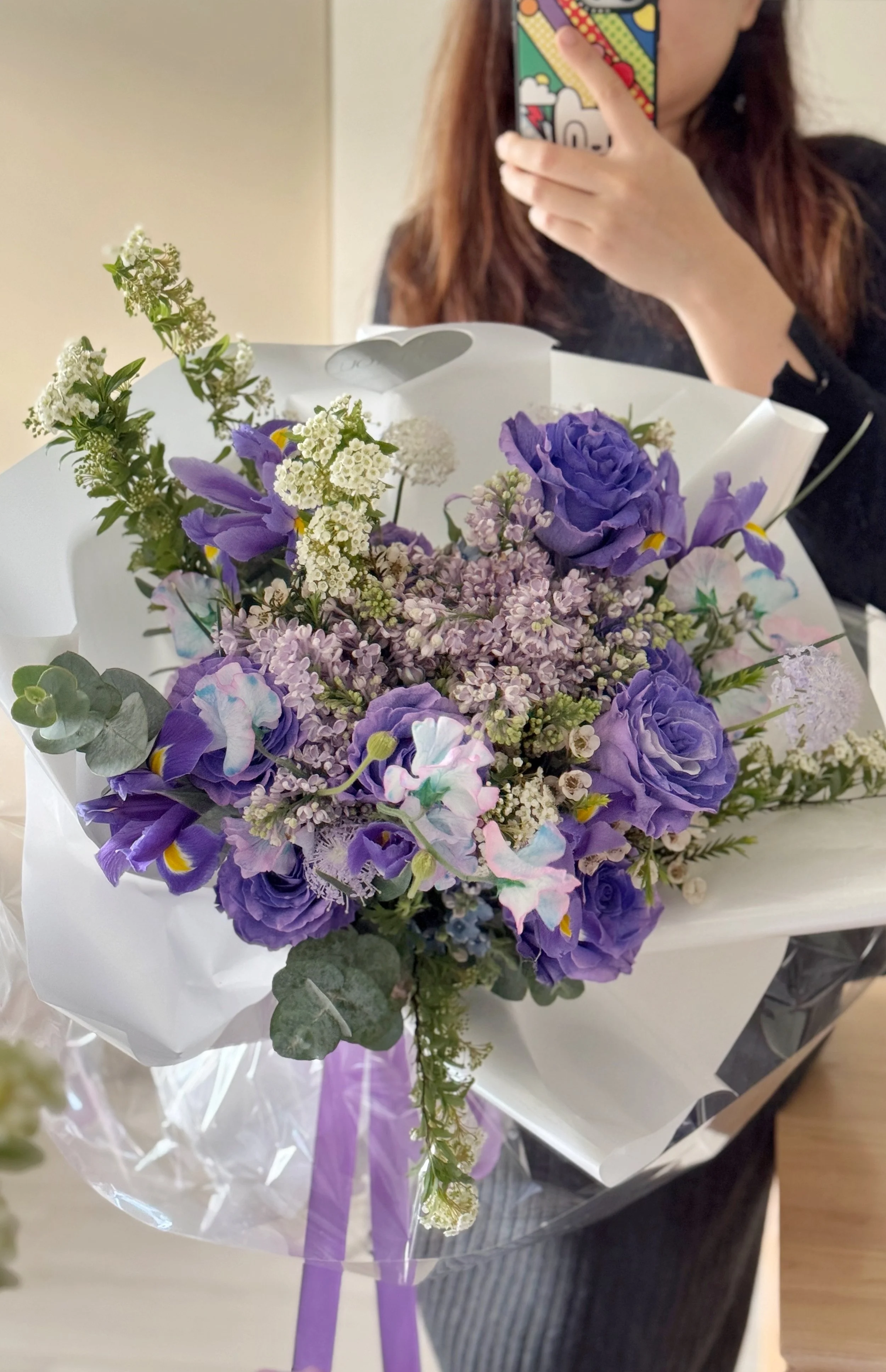 A person holding a bouquet of purple and white flowers, with a person taking a selfie in the background.