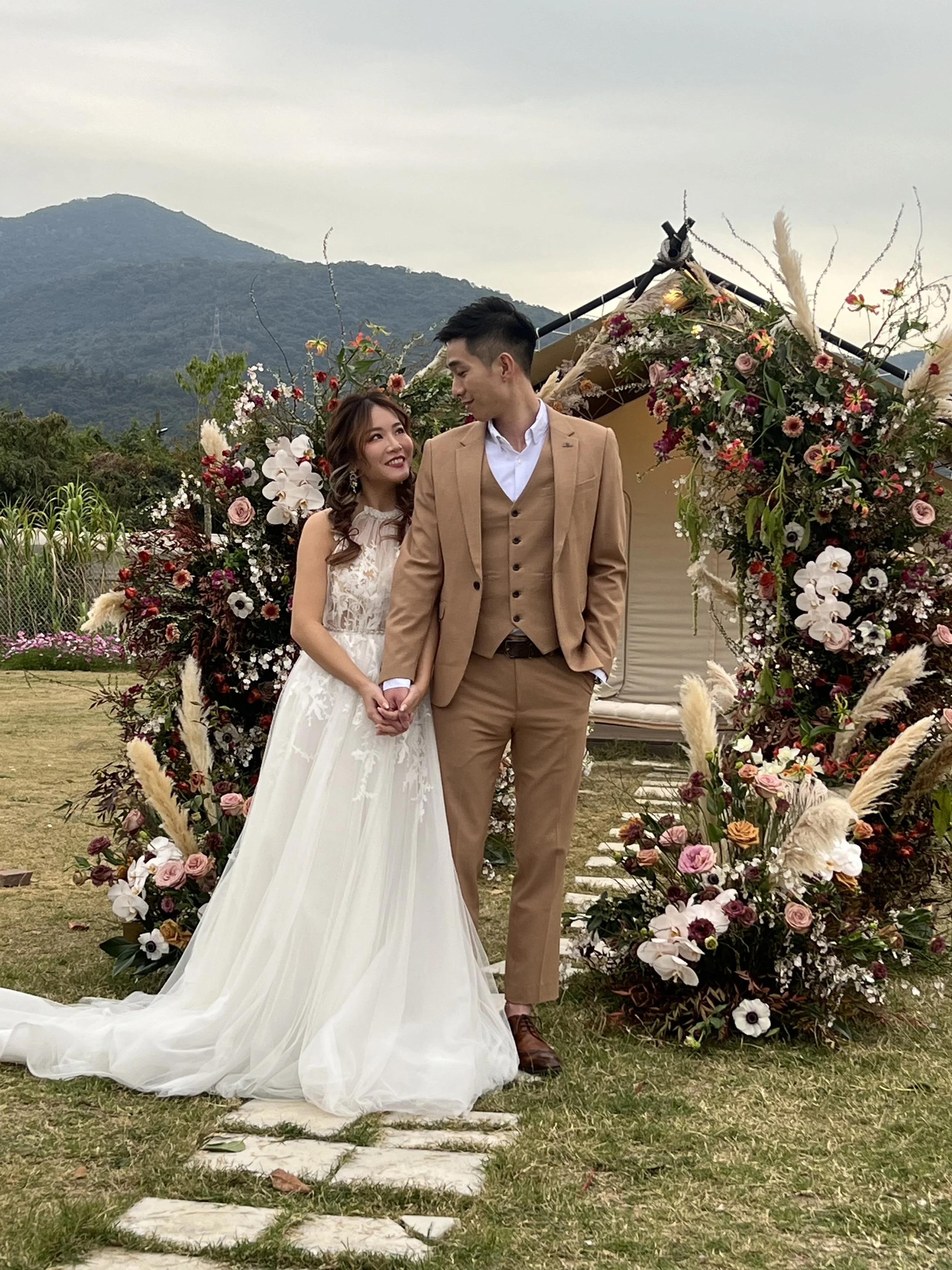 A bride and groom holding hands, standing on a stone pathway outside during a wedding ceremony, with mountains and a floral arch in the background.