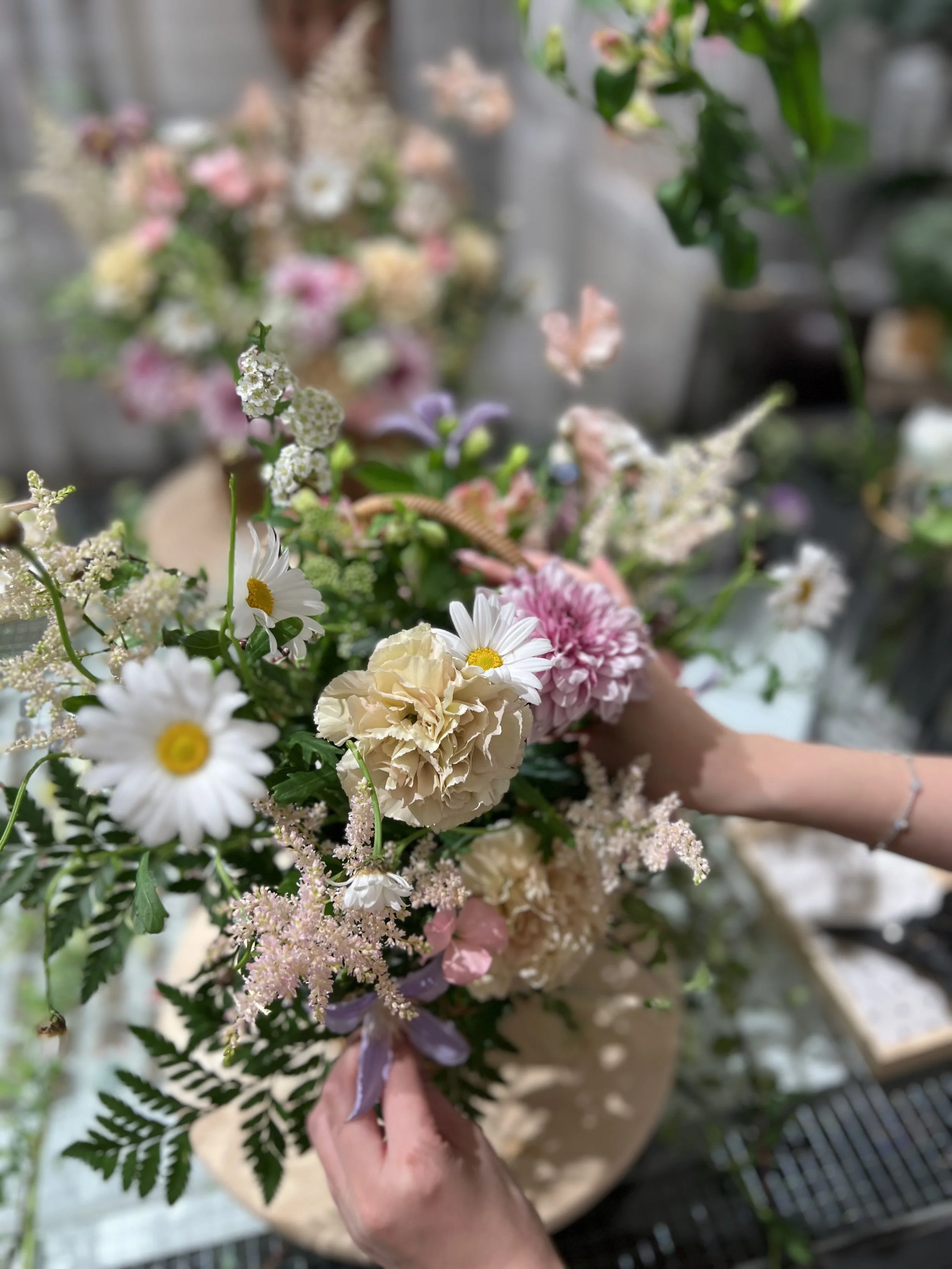 Close-up of a person holding a colorful bouquet of flowers with white daisies, cream roses, pink chrysanthemums, and other assorted flowers and greenery.