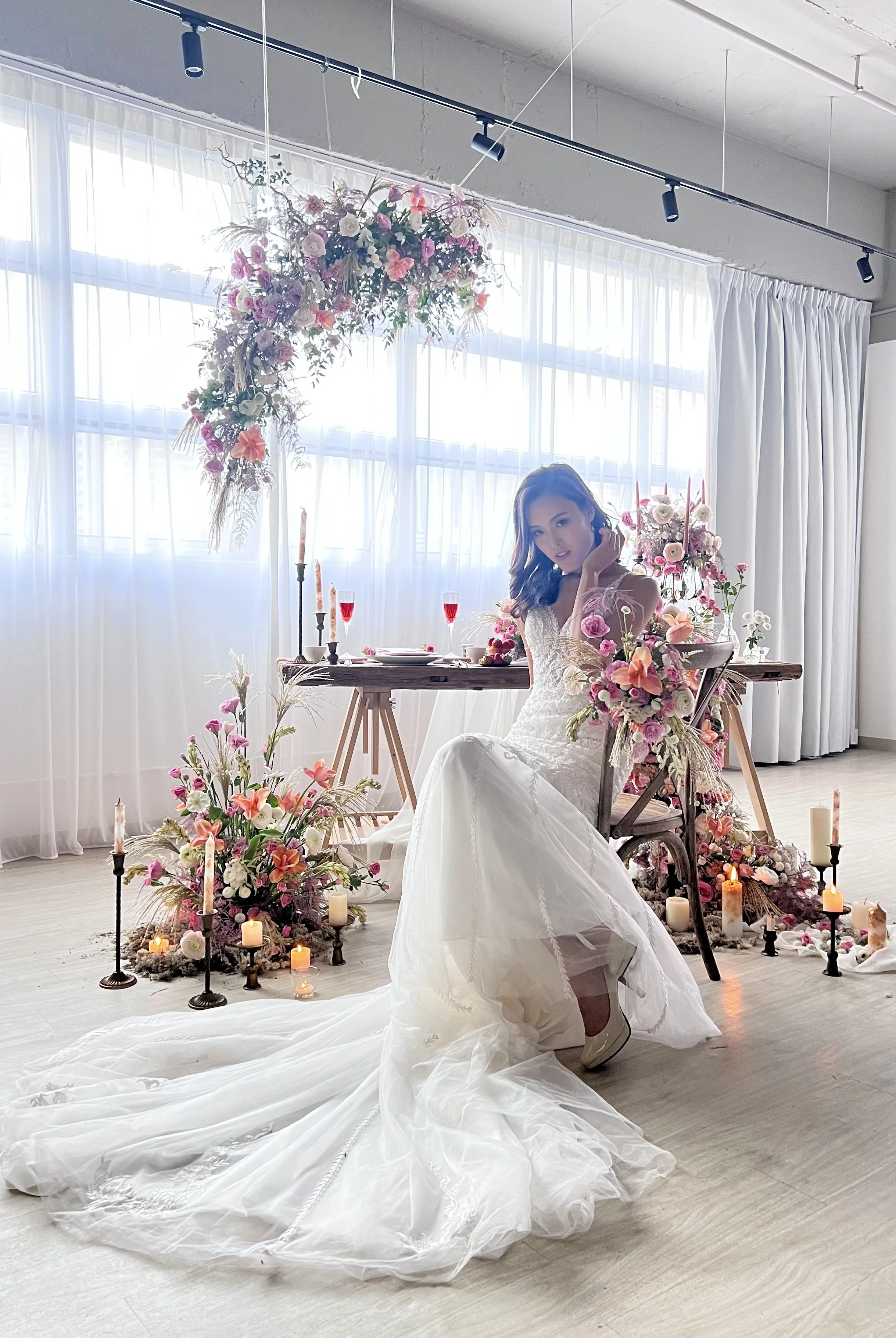 A woman in a wedding dress sitting at a decorated table with pink and white flowers, candles, and champagne flutes, in a bright room with white curtains.
