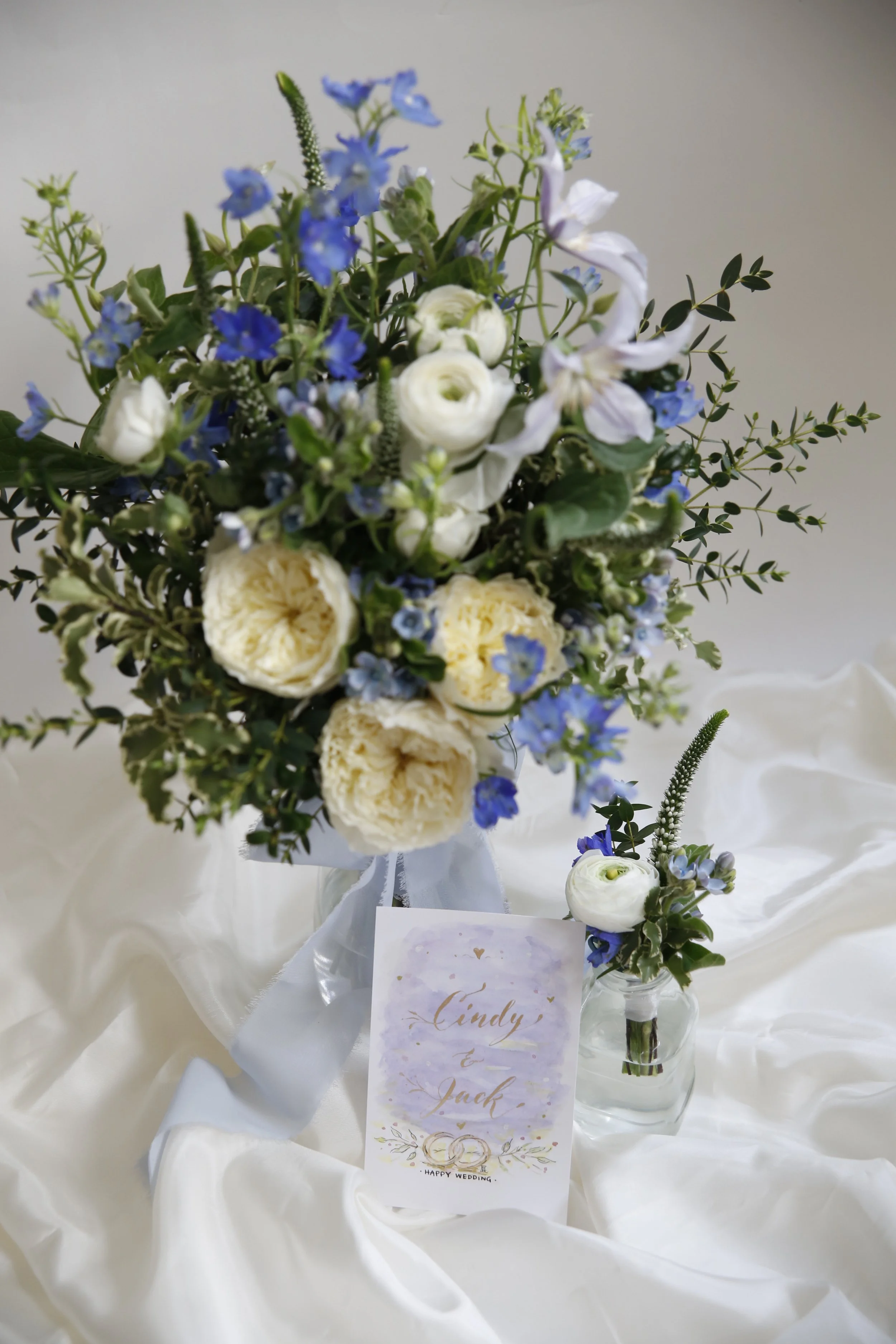 A wedding bouquet with white and blue flowers, and a personalized wedding card with the names Cindy and Jack, placed on satin fabric.