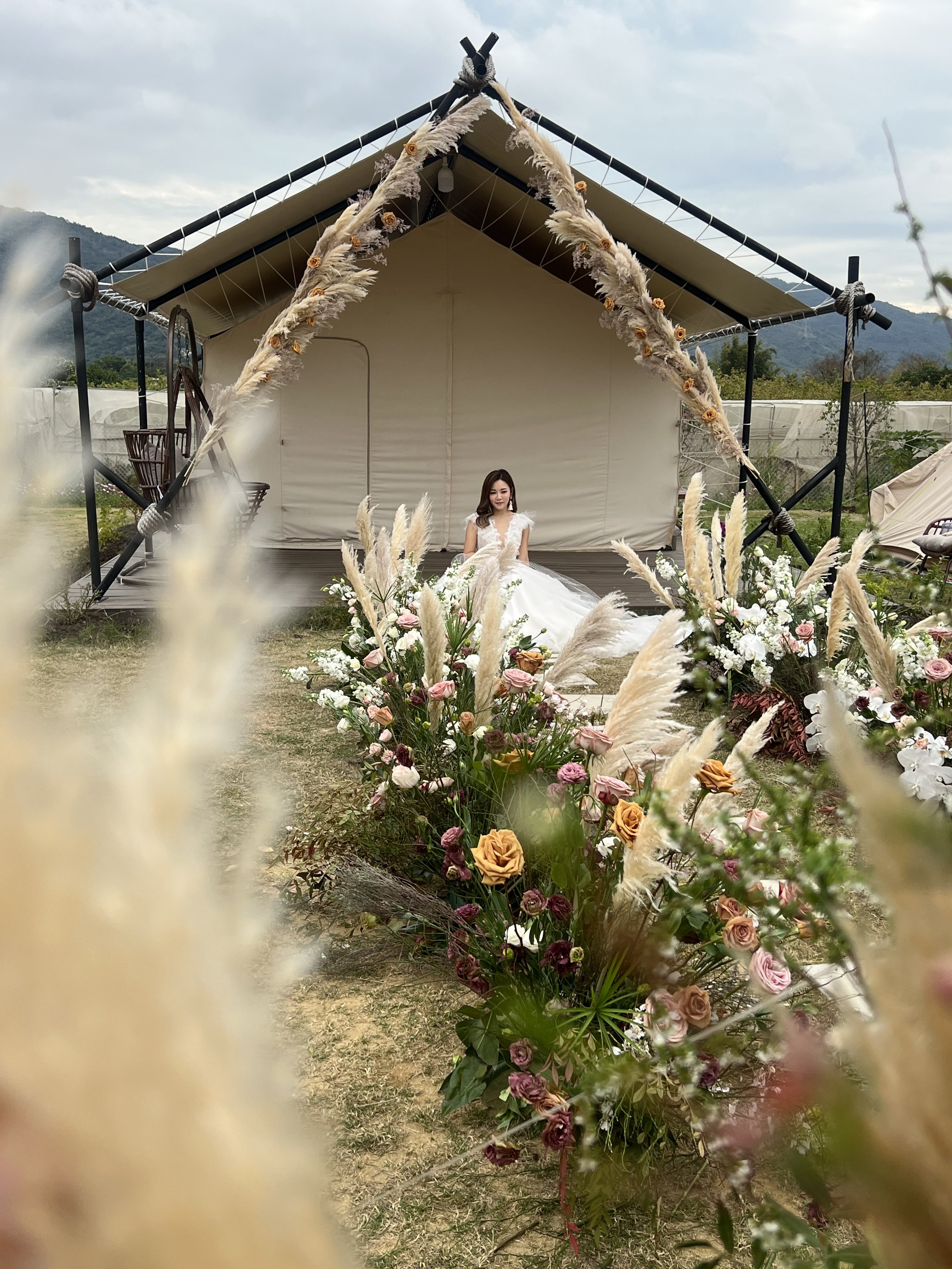 A bride in a white wedding gown sitting on the ground outdoors in front of a large tent decorated with pampas grass and flowers, with mountains and cloudy sky in the background.