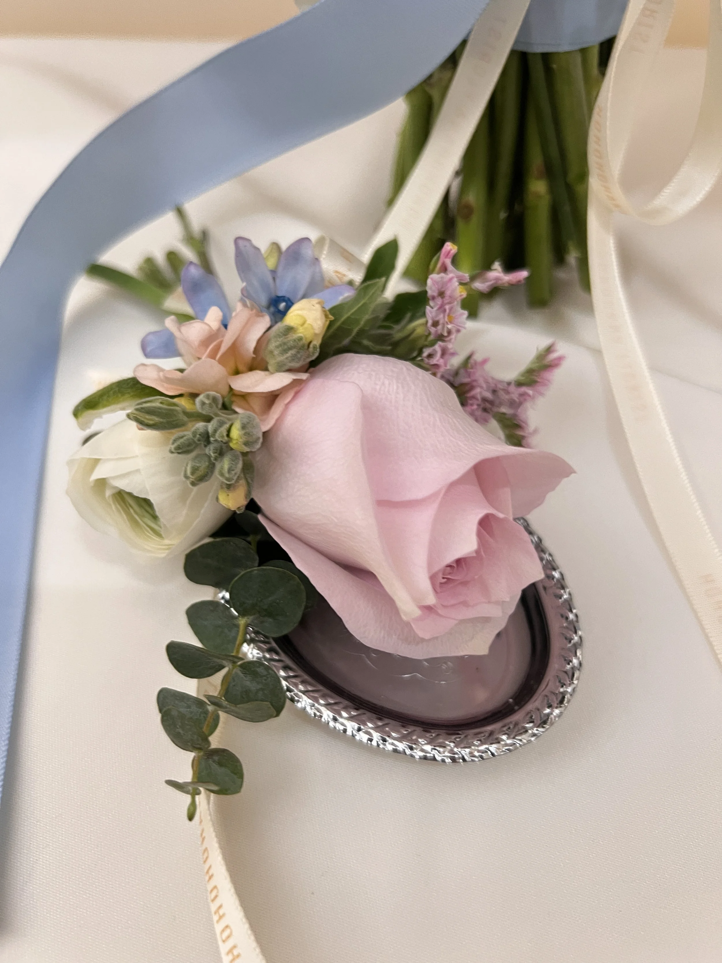 Close-up of a floral arrangement with a pink rose, light blue, white, and pink flowers, and greenery, resting on a silver tray on a white table with white and beige ribbons.
