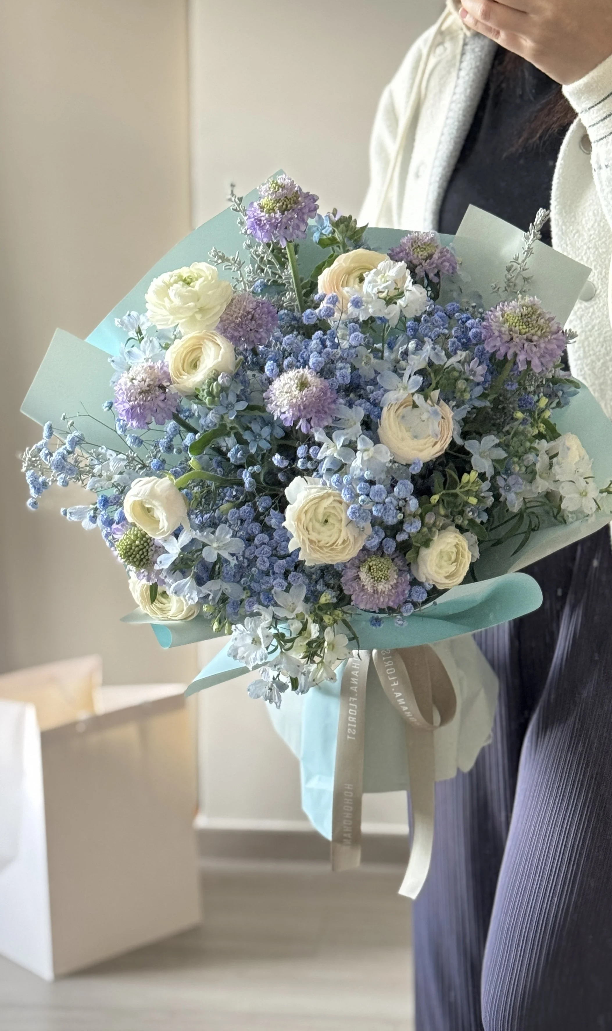 Person holding a bouquet of white, purple, and blue flowers wrapped in light blue paper, with a beige ribbon, indoors near a beige wall.