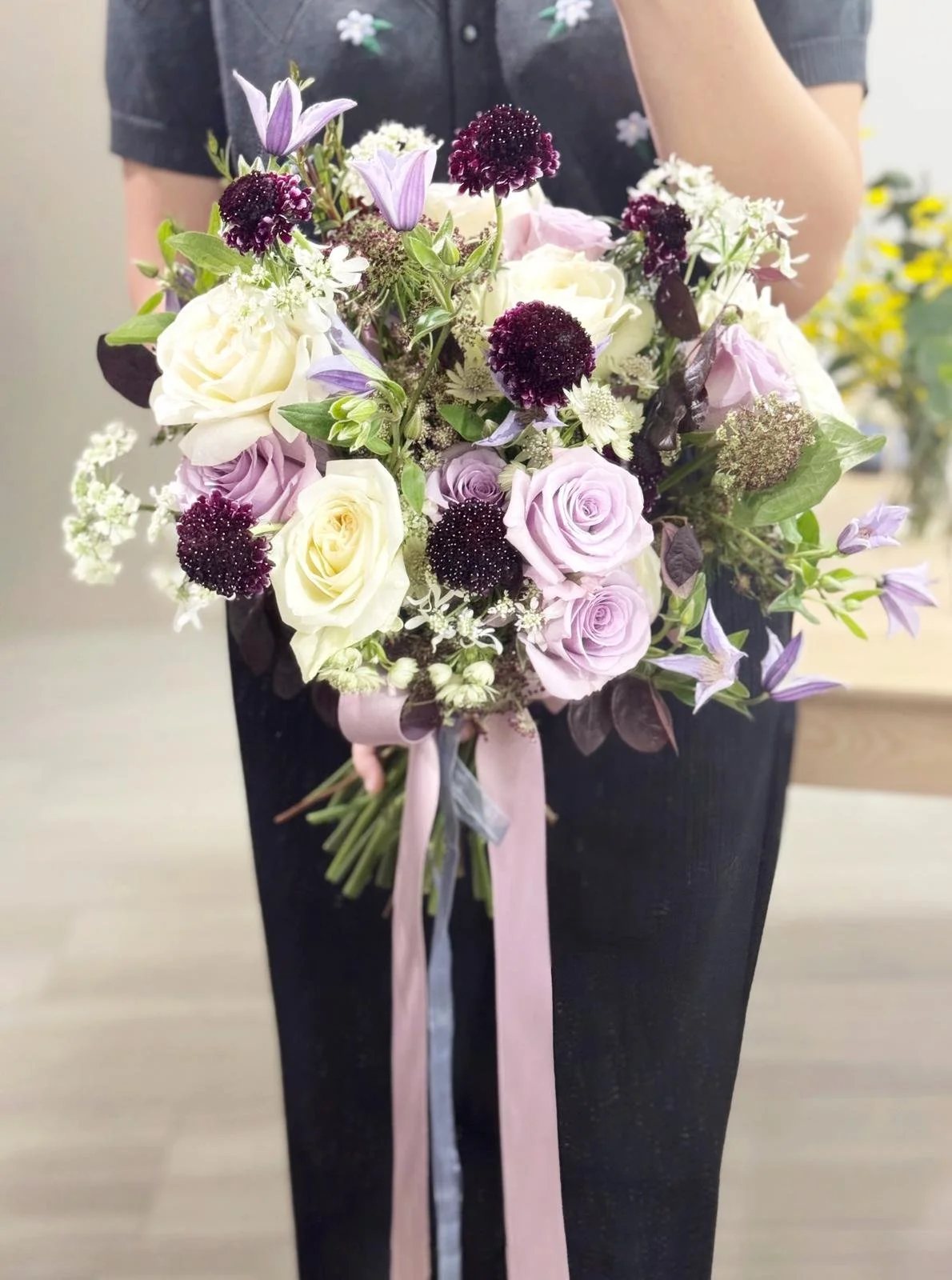 A person in a dark dress holds a bouquet of white and light purple roses, dark purple billy buttons, and other small flowers tied with pink and silver ribbons.
