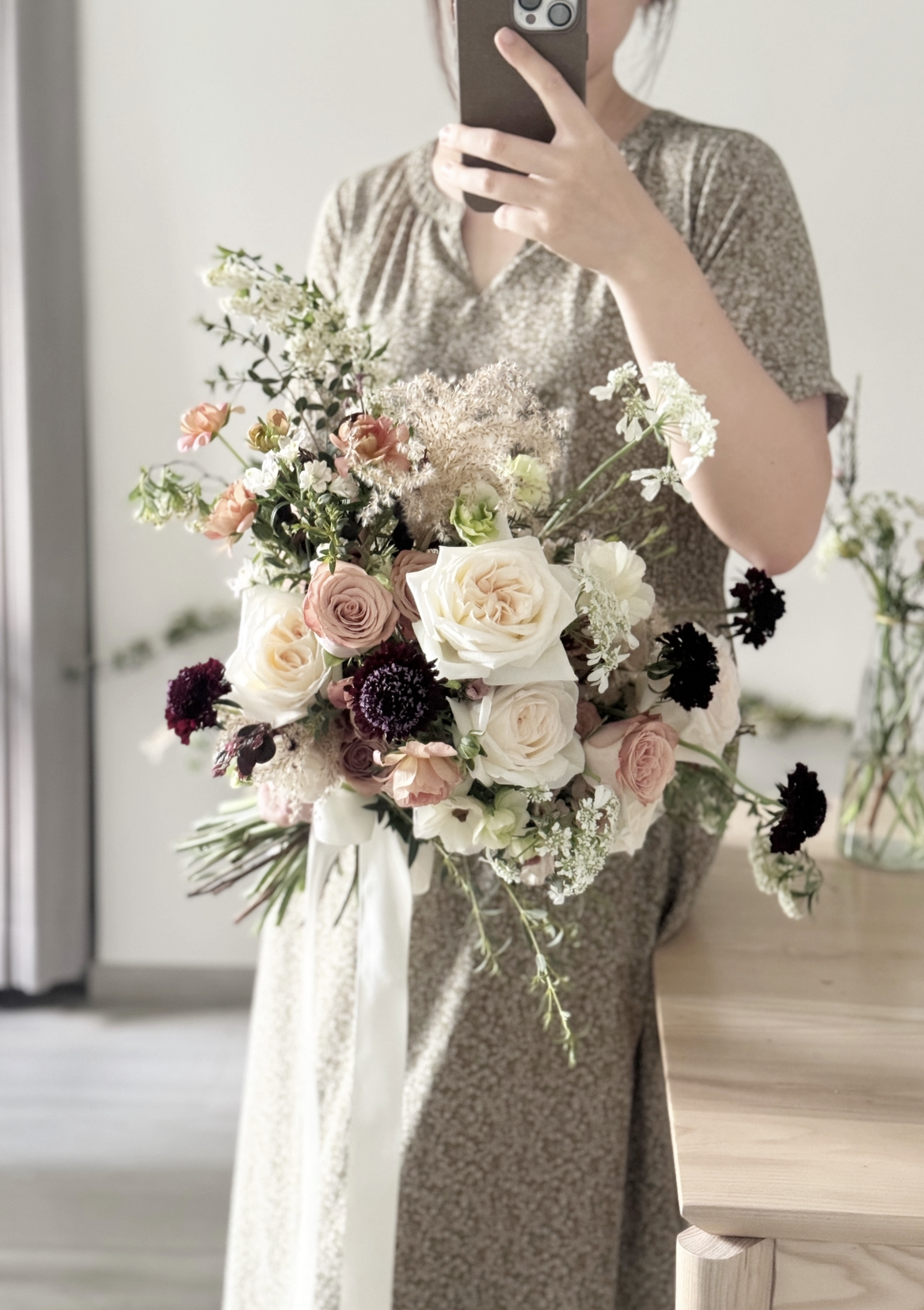 Person taking a mirror selfie holding a bouquet of flowers with cream, pink, and dark purple blooms, in a light-colored dress.