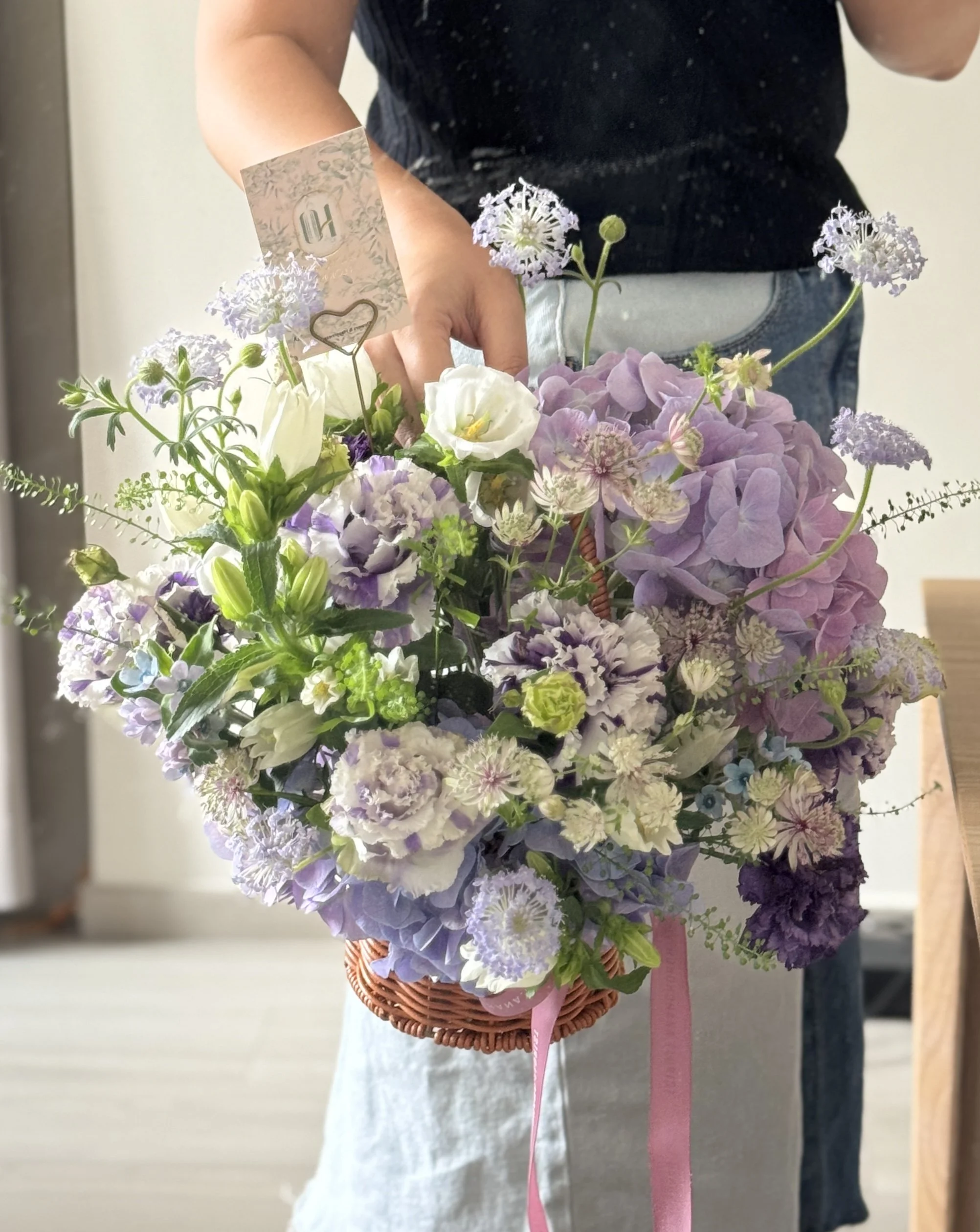 Person holding a basket of purple, white, and green flowers, with a gift card attached to the arrangement.