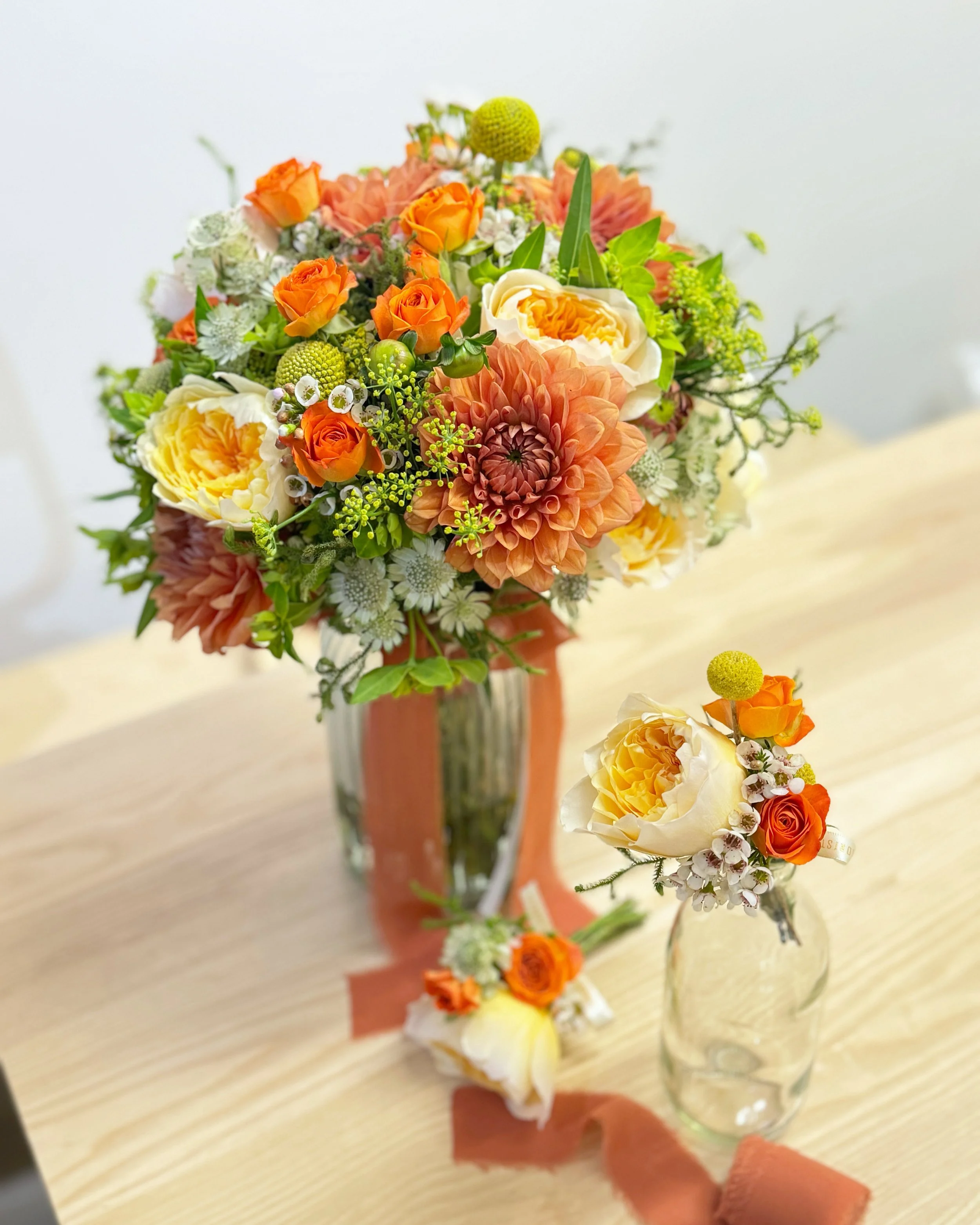 Colorful flower arrangements with orange, white, and yellow roses, peach dahlias, and green accents in glass vases on a wooden table.